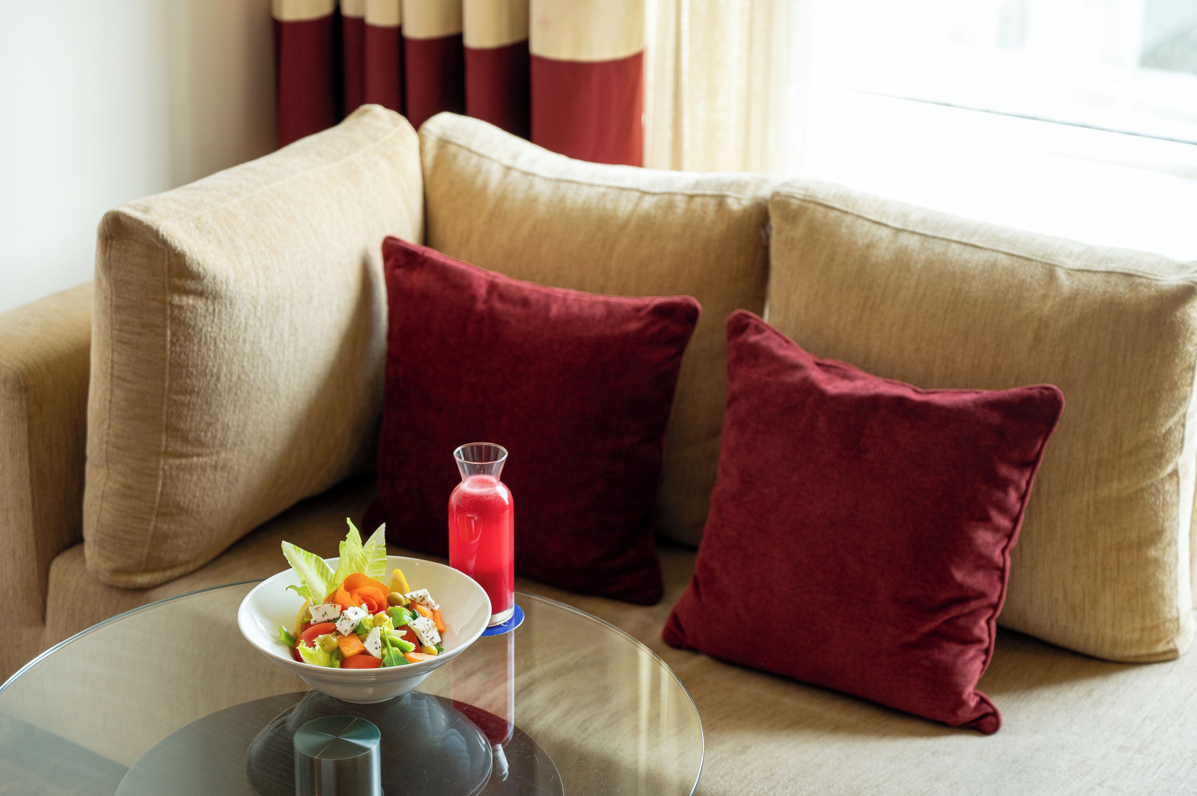 Een glazen tafel met een saladeschaal en een glas rode drank op een beige bank met rode kussens.