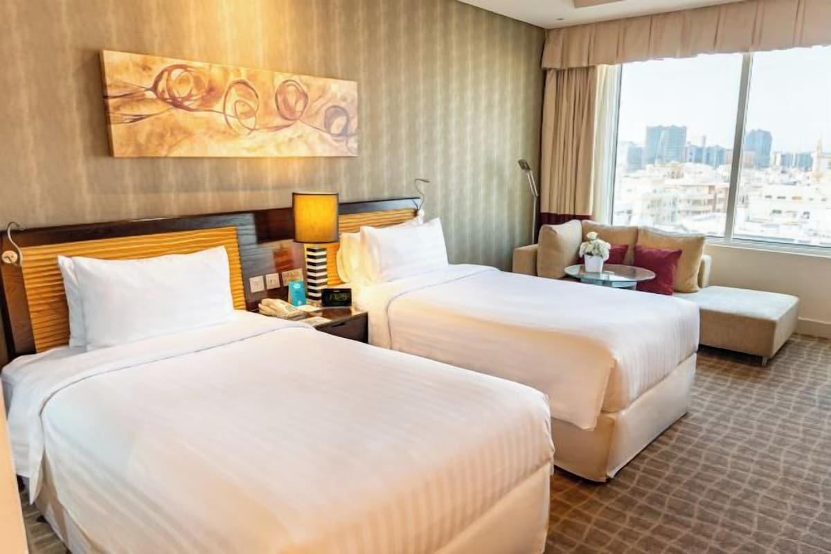 A hotel room with two beds, a sofa, a city view, and a painting on the wall.