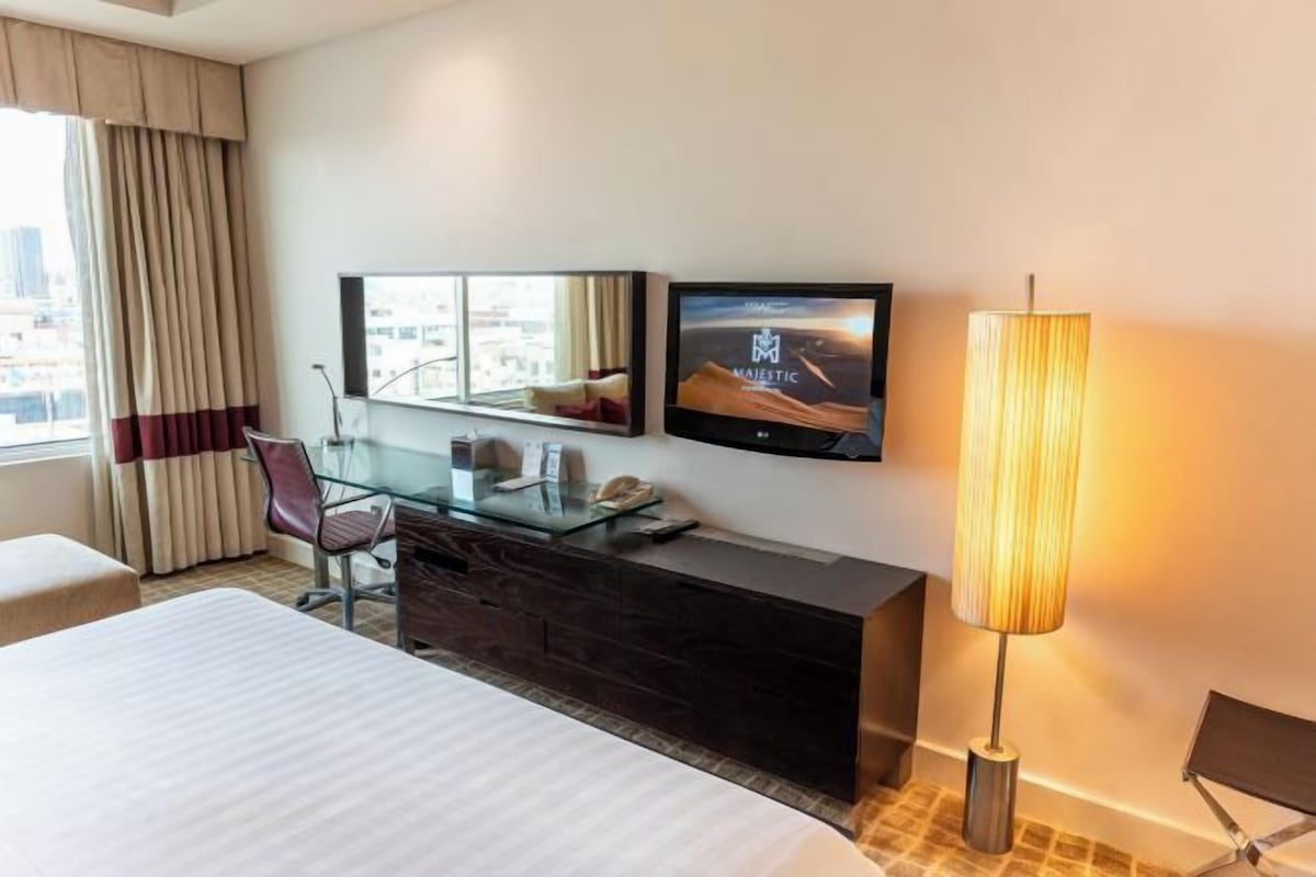 A hotel room with a bed, a desk, a chair, a TV, a lamp, and a window with curtains.