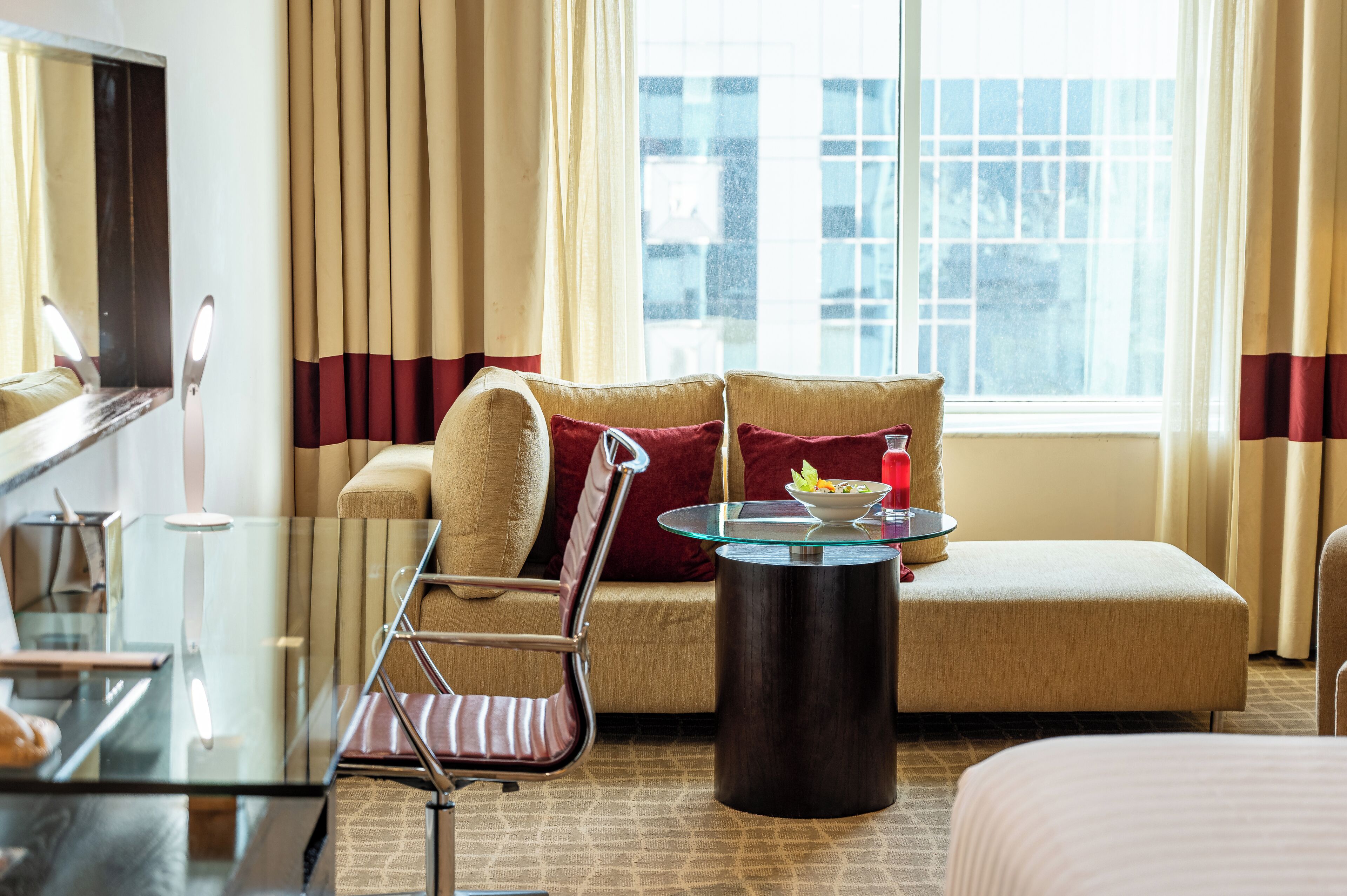 Habitación de hotel con sofá, una mesa de centro de vidrio, una silla, una cama y una ventana con cortinas.