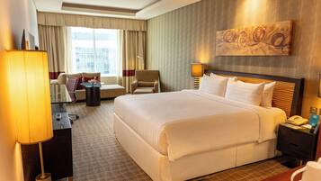 A hotel room with a large bed, a desk with a lamp, a chair, a sofa, and a painting on the wall.