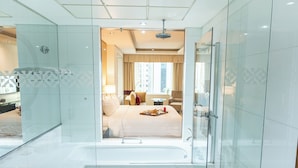 A modern bathroom with a glass-enclosed shower, a bathtub, and a visible bedroom with a bed and a nightstand.
