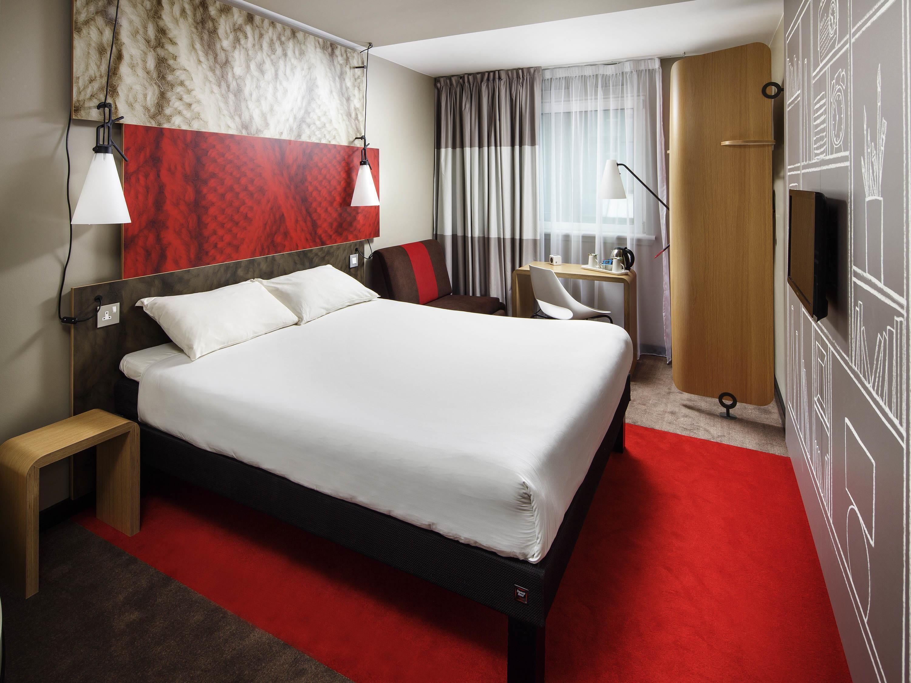 A hotel room with a large bed, a desk, a chair, a TV, and a red carpet.
