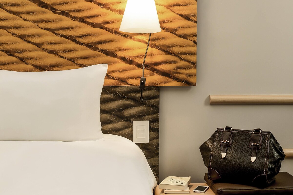 Una habitación de hotel con una cama, una mesita de noche con una lámpara, una luz empotrada en la pared, un bolso de mano y un teléfono móvil.