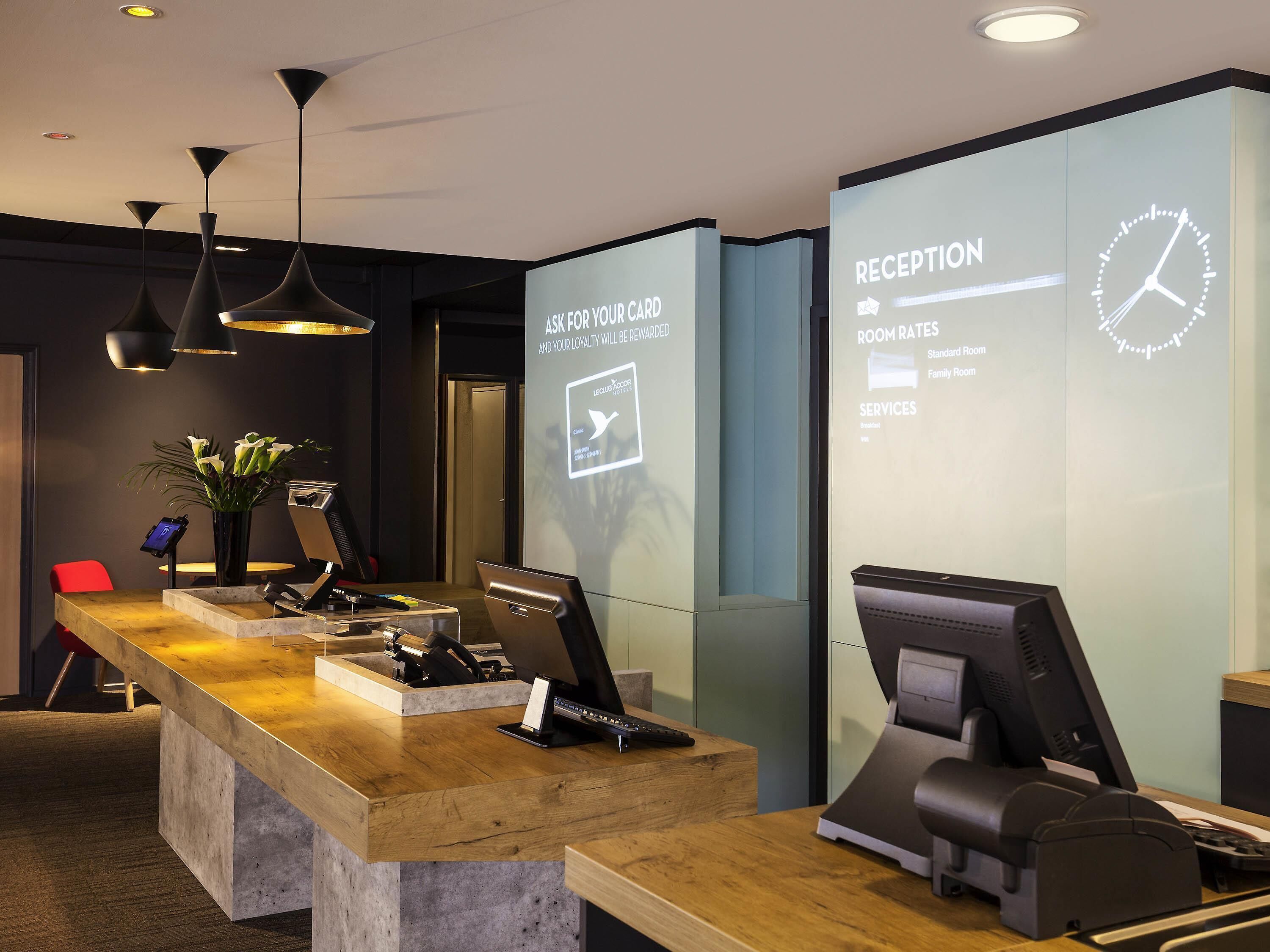 Een receptiebalie van een hotel met een digitaal scherm dat prijzen en diensten toont, een houten balie en een modern plafondontwerp.
