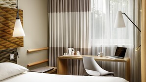 Une chambre d’hôtel moderne, équipée d’un lit, d’un bureau avec un ordinateur portable, d’une chaise, d’une lampe et d’une fenêtre avec des rideaux.