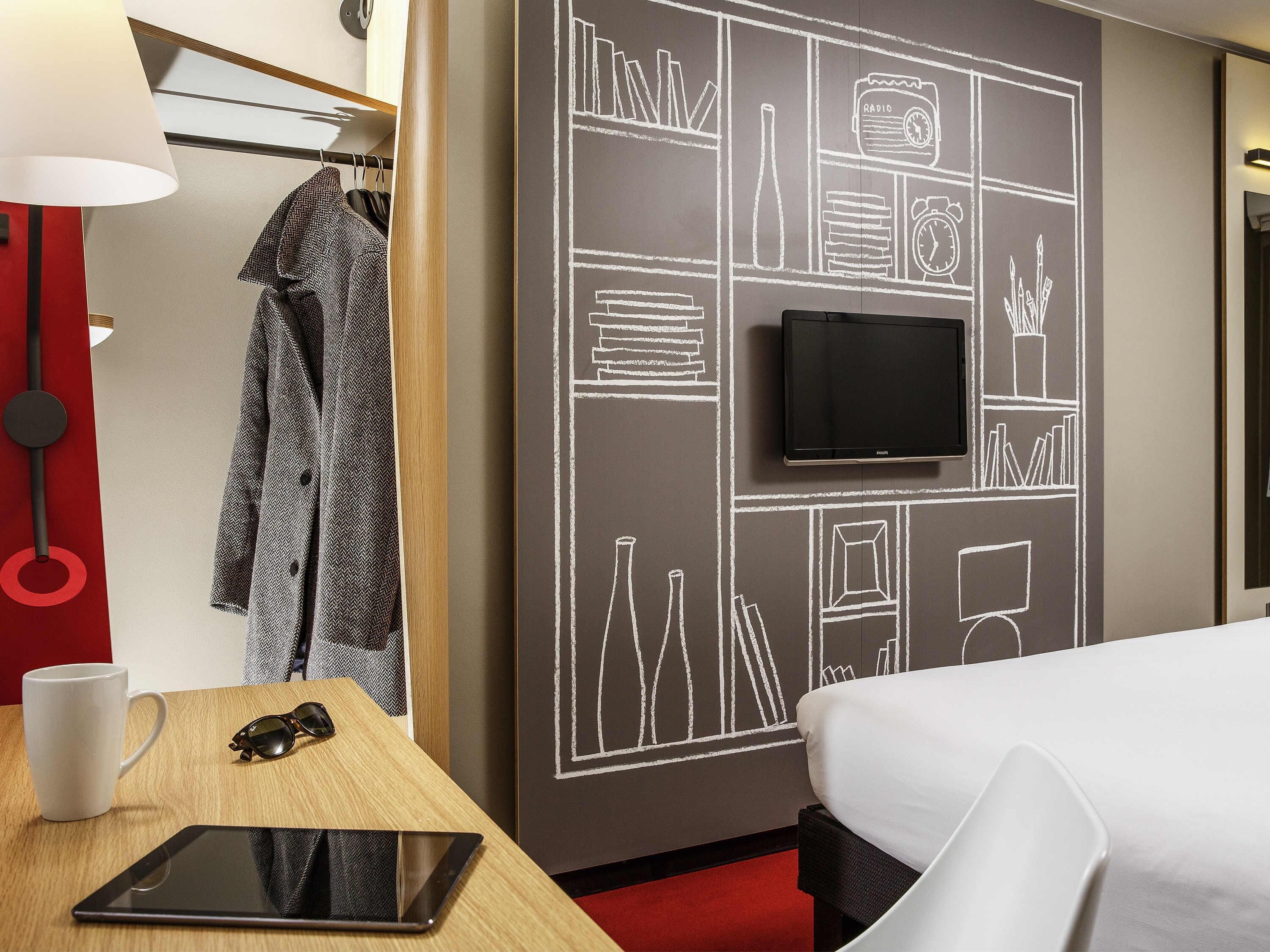 Una habitación de hotel con una cama, un escritorio con una tableta, un televisor y una pared con diseño de pizarra.