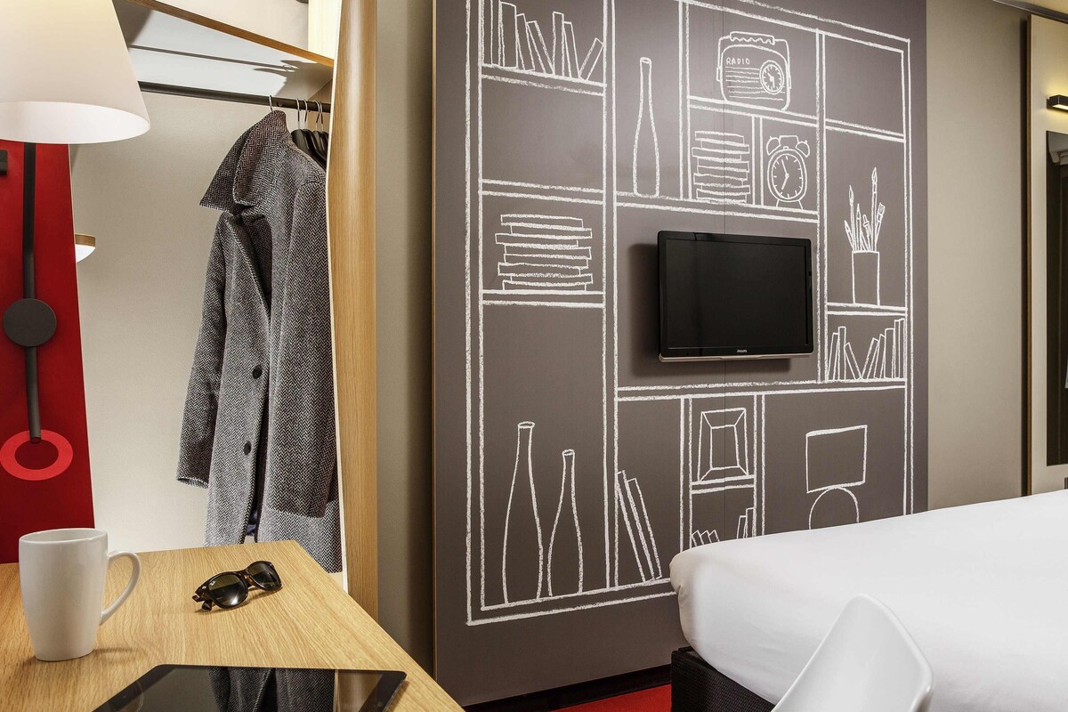 Una habitación de hotel con una cama, un escritorio con una tableta, un televisor y una pared con diseño de pizarra.