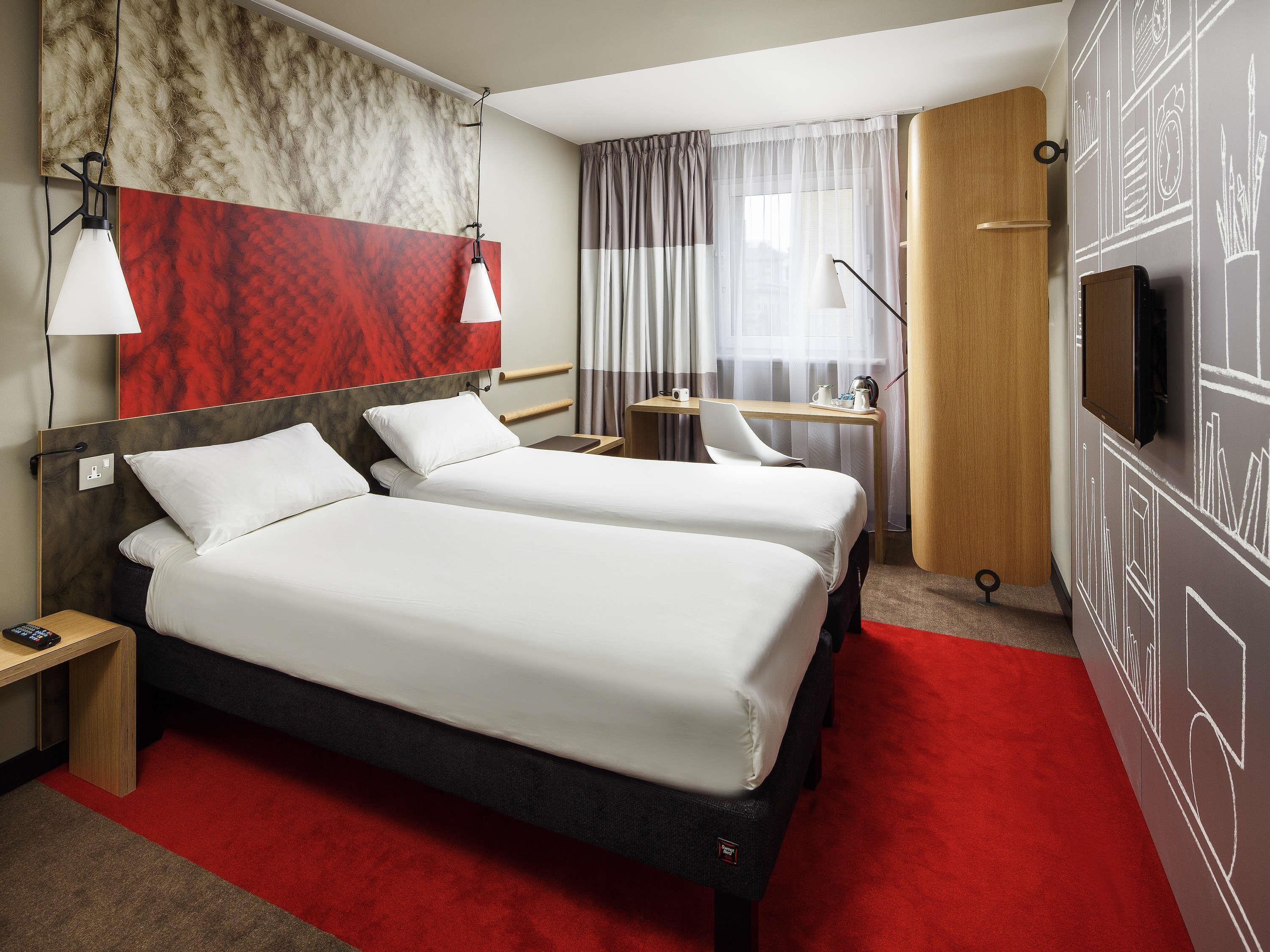 A hotel room with two beds, a desk, a TV, and a red carpet.