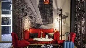 Une chambre d’hôtel moderne avec un canapé rouge, une petite table ronde ornée d’un vase et d’une tasse, une valise bleue et une grande fresque murale.