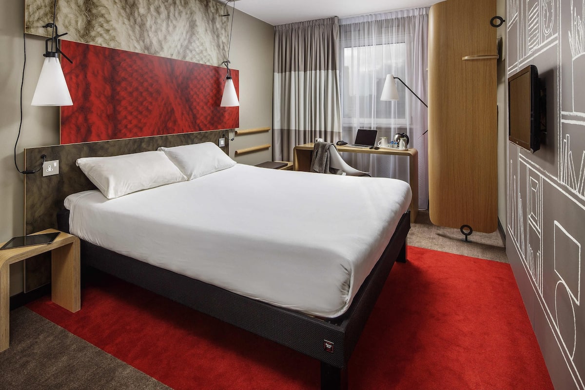 A hotel room with a bed, bedside table, desk with a laptop, and a TV on the wall.