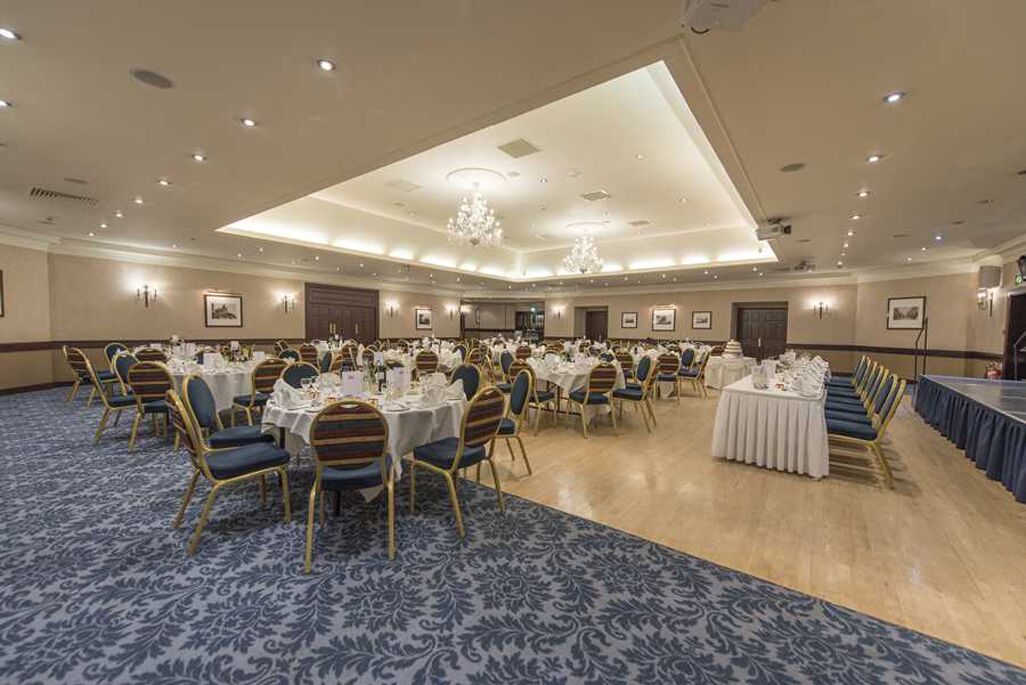 Un amplio salón de banquetes con mesas redondas y sillas, una alfombra estampada y un techo alto con iluminación empotrada.