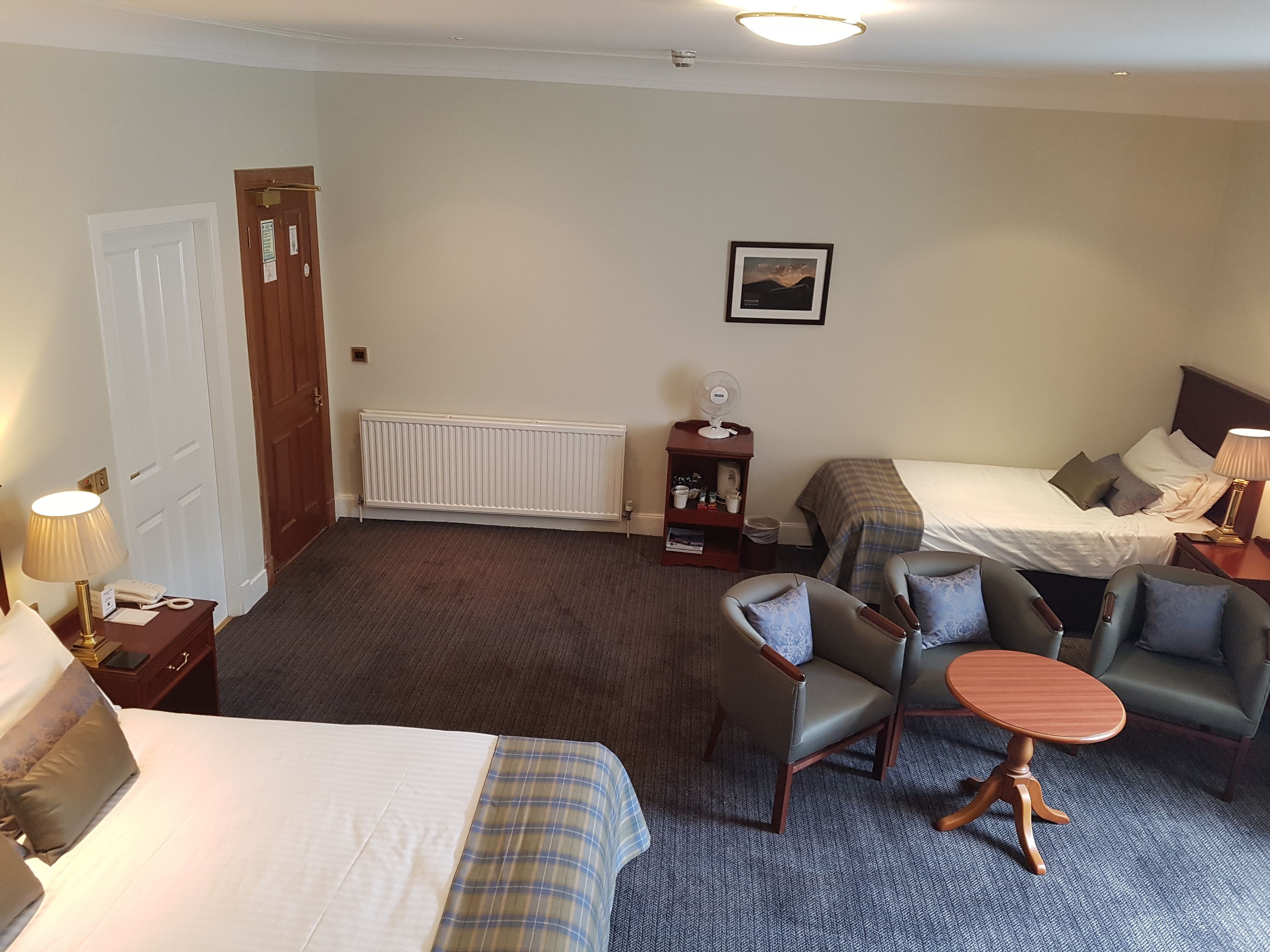 A hotel room with two beds, a sitting area with chairs, a small table, a radiator, a bedside table with a lamp, a picture on the wall, and a door.