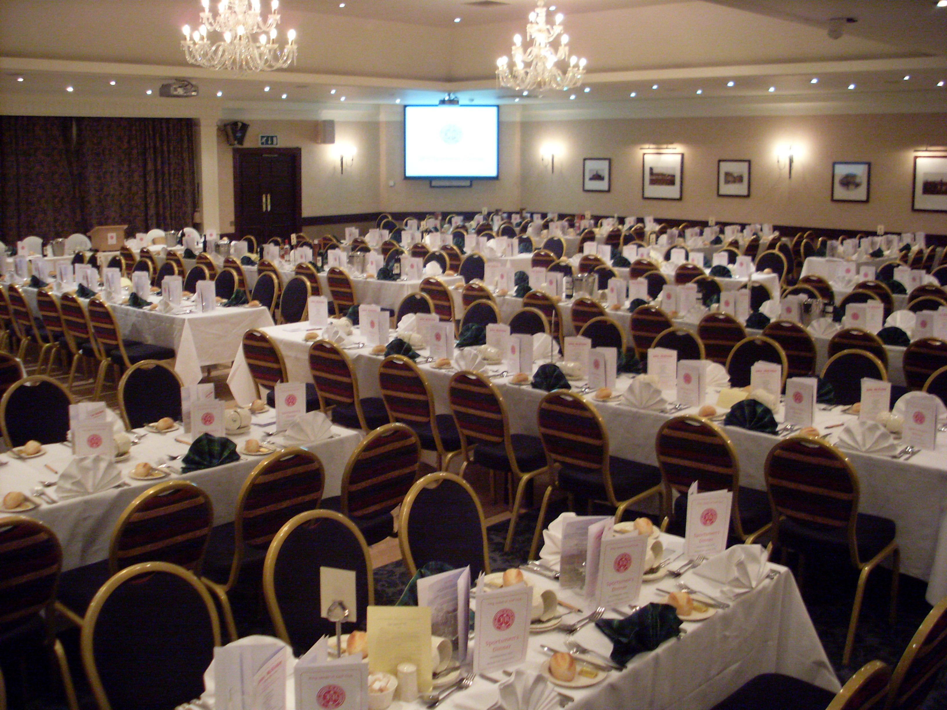 A banquet hall with rows of tables set for a formal event, featuring gold chairs and white tablecloths.