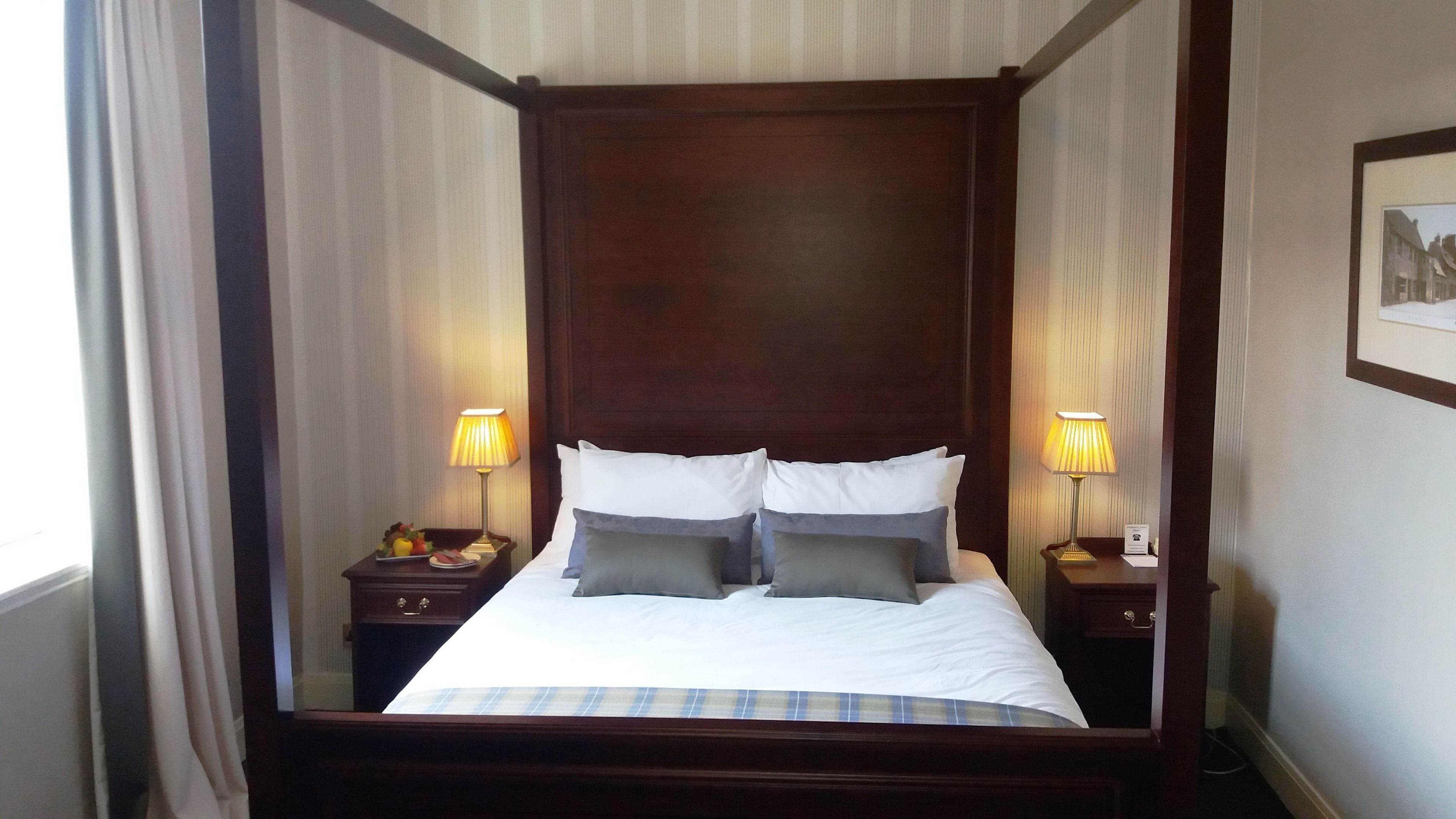 Una cama con dosel con sábanas blancas y almohadas grises, flanqueada por dos mesitas de noche con lámparas, en una habitación con papel tapiz a rayas.