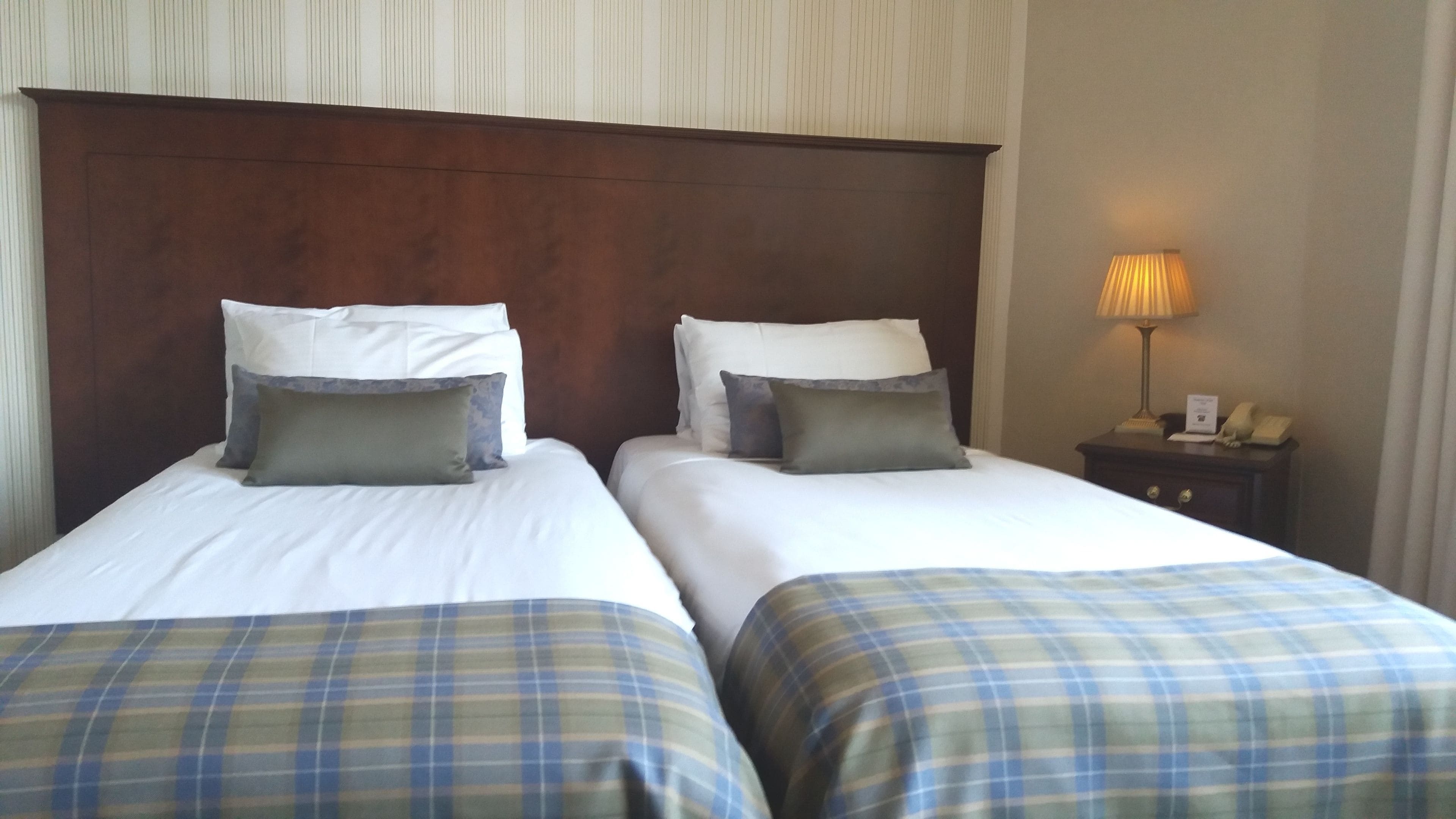 Habitación de hotel con dos camas, cada una con sábanas blancas y colchas a cuadros. Tiene un cabecero de madera, una mesita de noche con lámpara y teléfono.