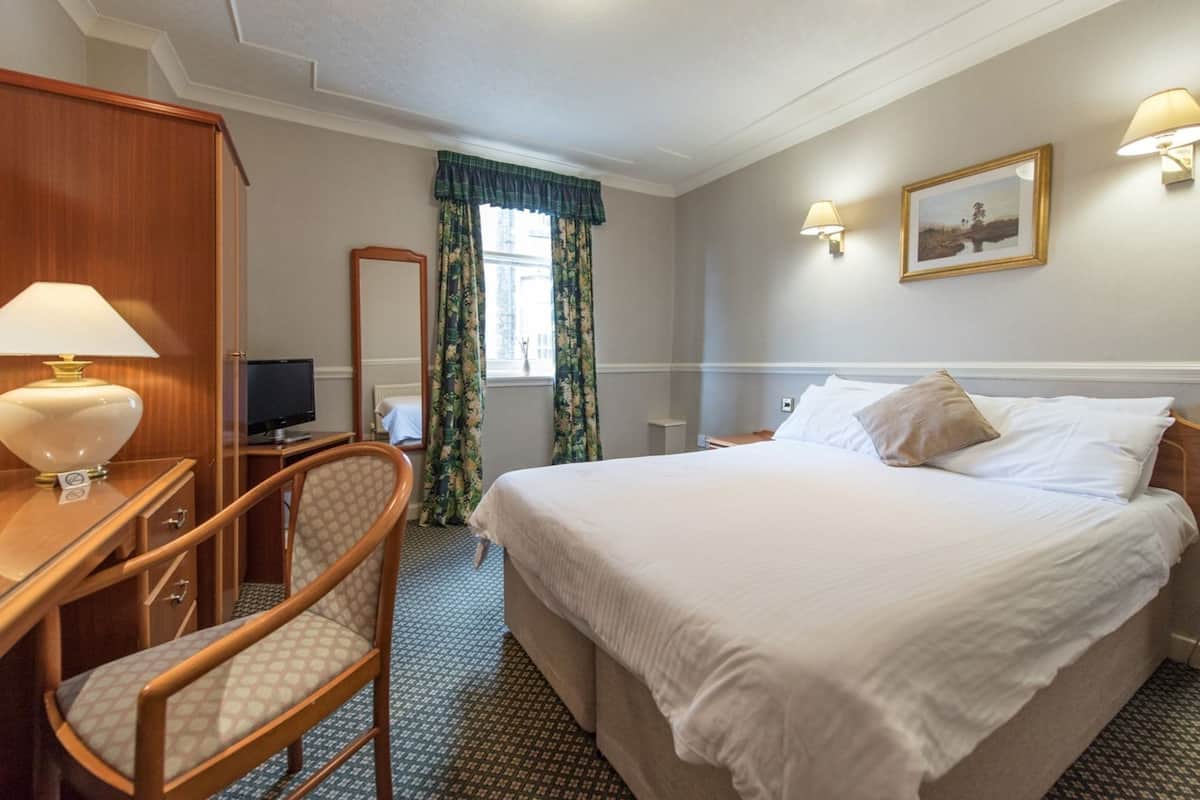A hotel room with a bed, a desk with a lamp, a chair, a television, a window with curtains, and wall-mounted lights.