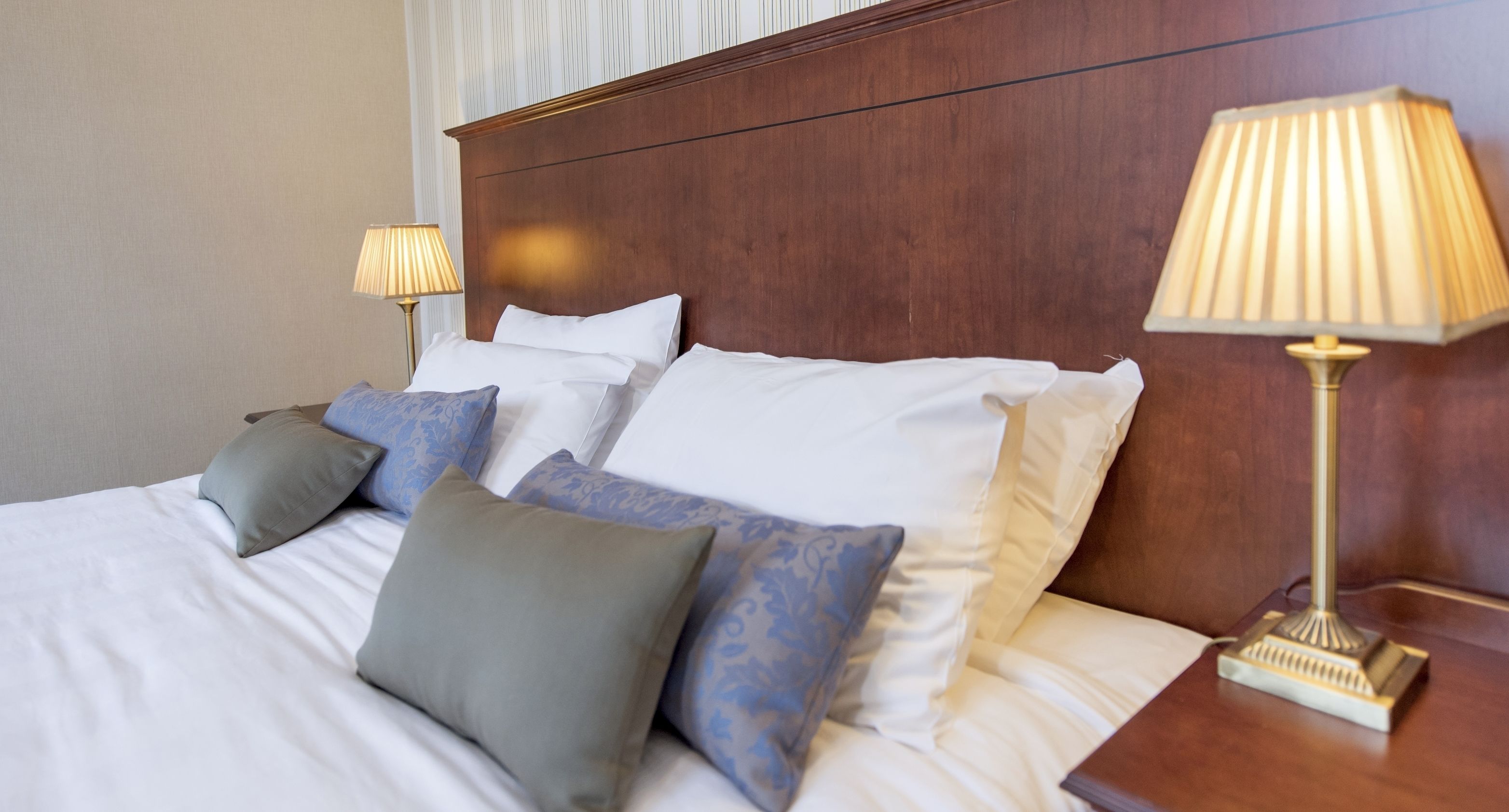A neatly made bed with white and grey linens, a wooden headboard, and two bedside lamps.