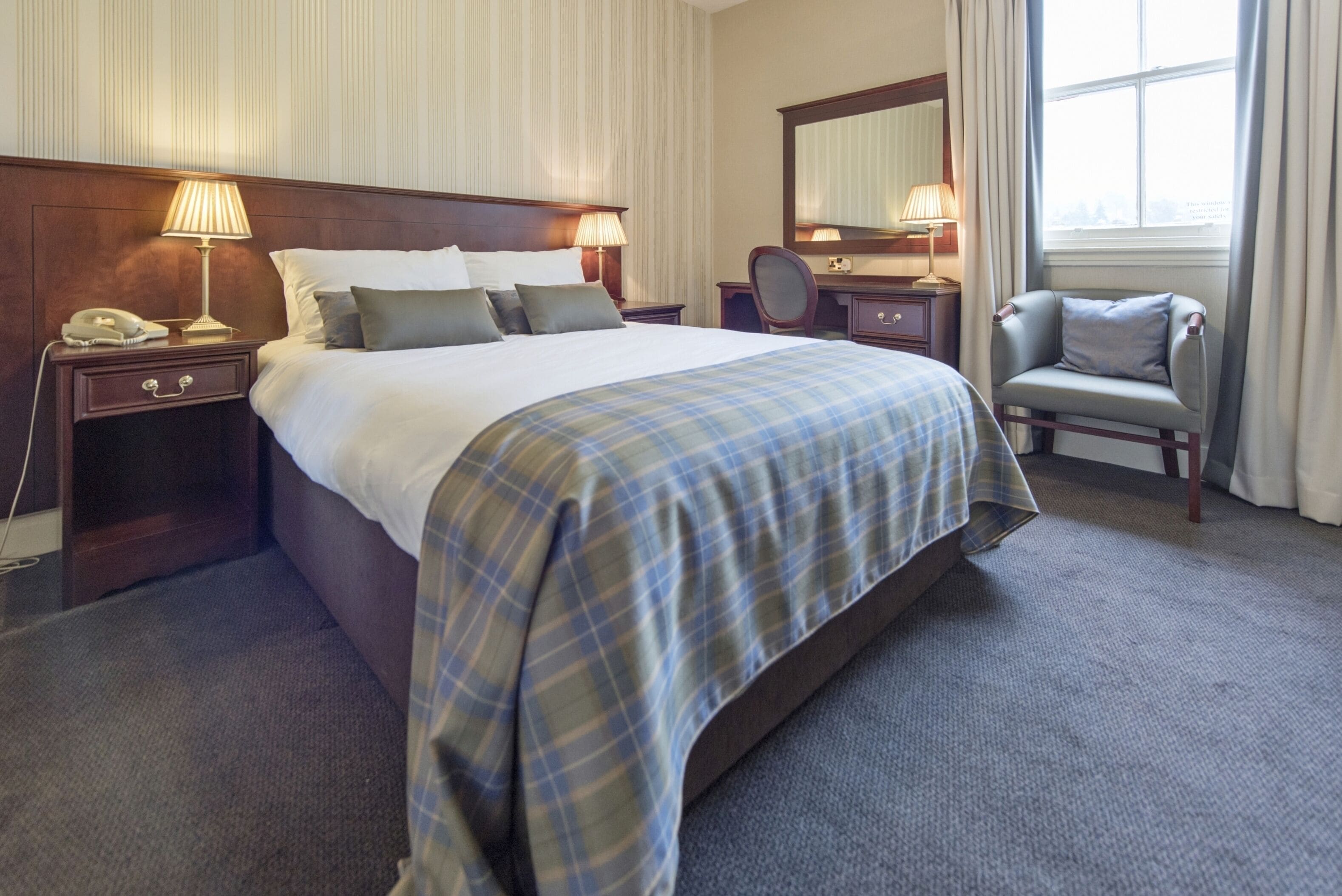 A hotel room with a large bed, bedside tables with lamps, a chair, a desk with a mirror, and a window with curtains.
