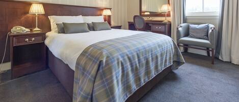 A hotel room with a large bed, bedside tables with lamps, a chair, a desk with a mirror, and a window with curtains.