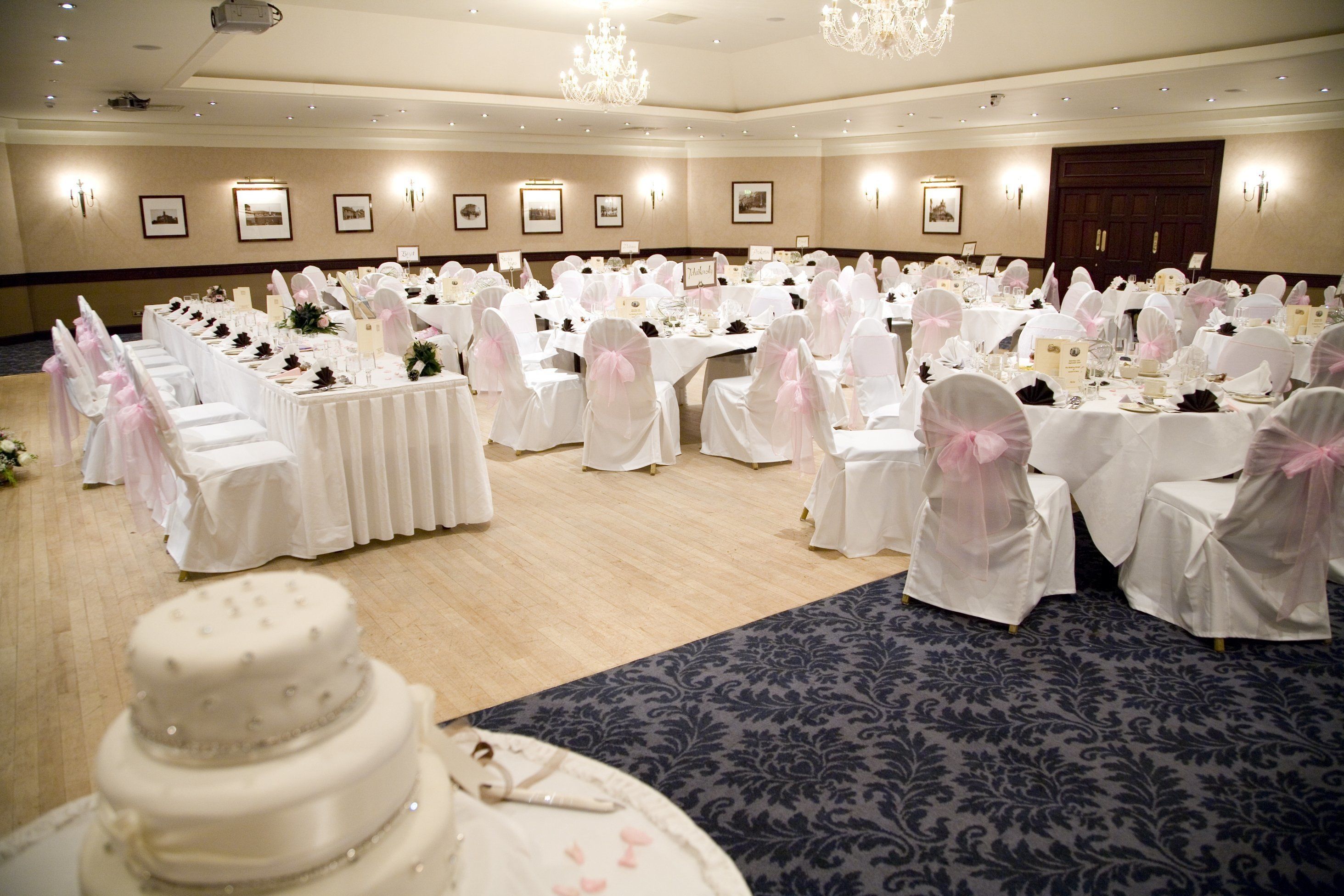 Un salón de banquetes con mesas redondas cubiertas con manteles blancos y sillas adornadas con cintas rosas, dispuesto para un evento formal.