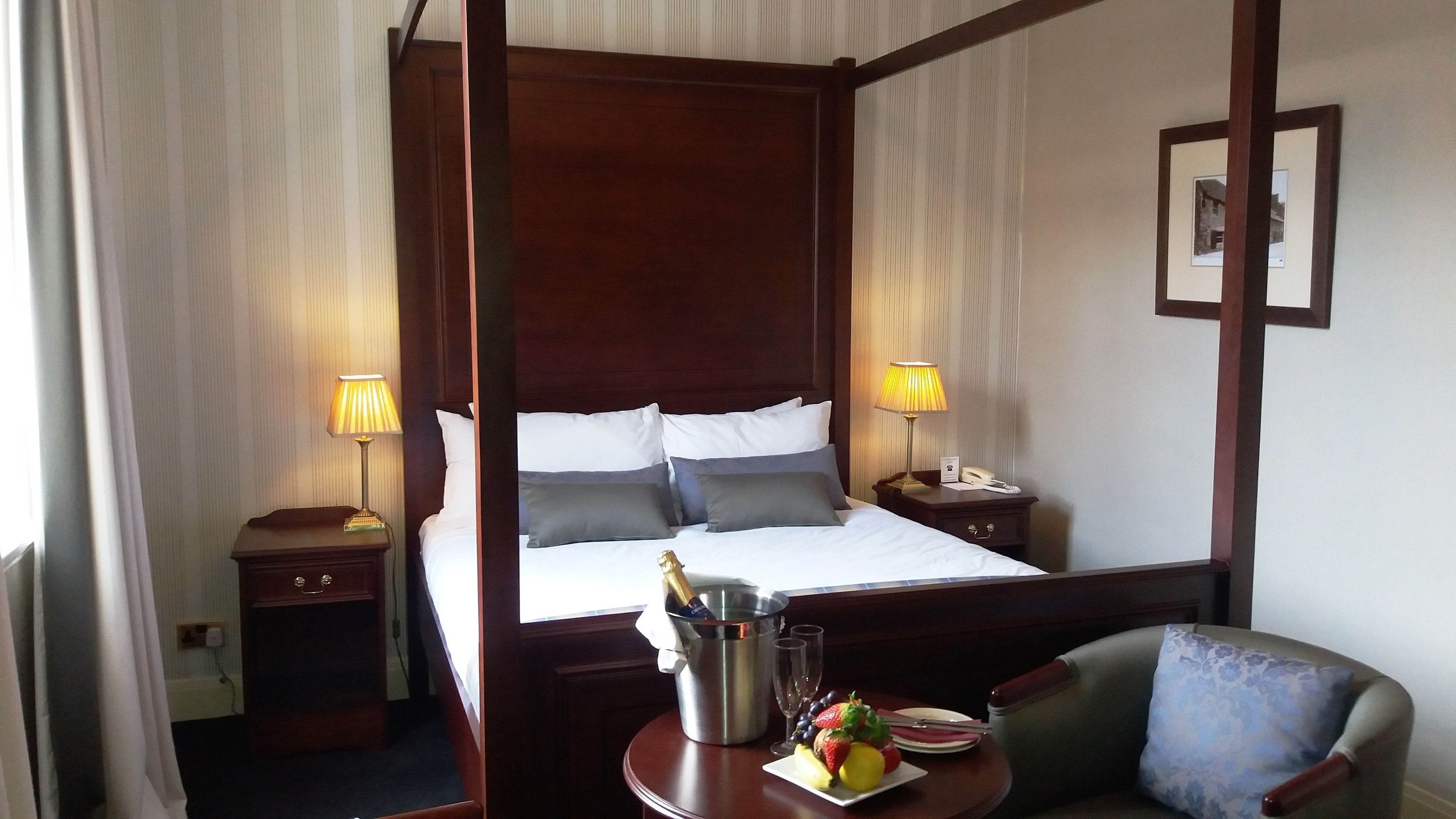 A four-poster bed with white and gray linens, two bedside lamps, a minibar with a bottle of champagne, and a fruit plate on a small table.