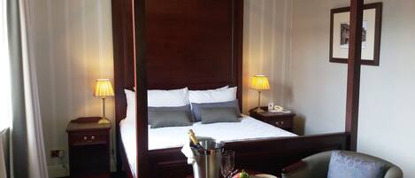 A four-poster bed with white and gray linens, two bedside lamps, a minibar with a bottle of champagne, and a fruit plate on a small table.