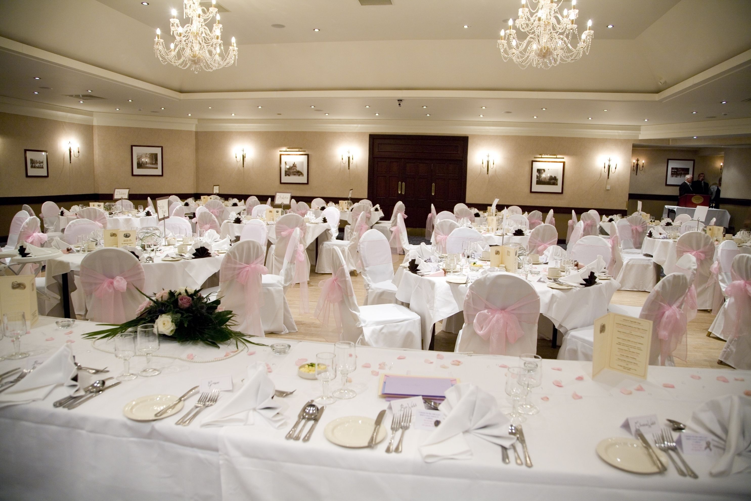 A banquet hall with round tables set for a formal event, featuring white tablecloths, pink sashes, and floral centerpieces.