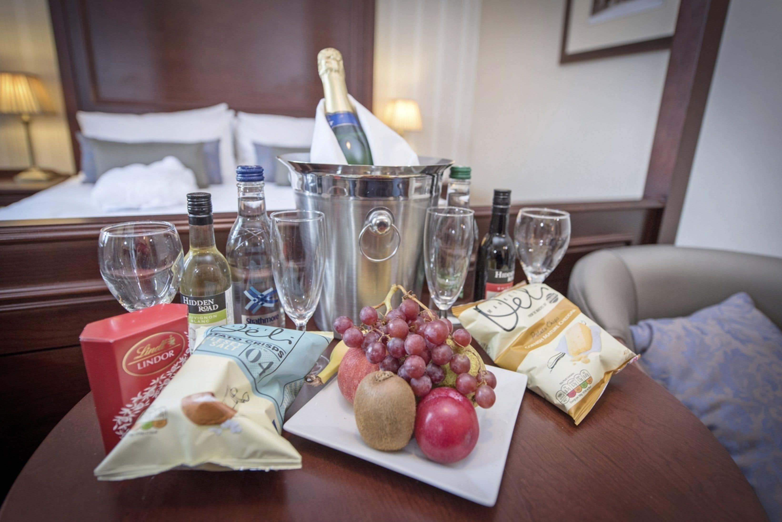 Une chambre d’hôtel avec un lit, une table de chevet sur laquelle se trouve une bouteille de champagne, des verres à vin et des en-cas.