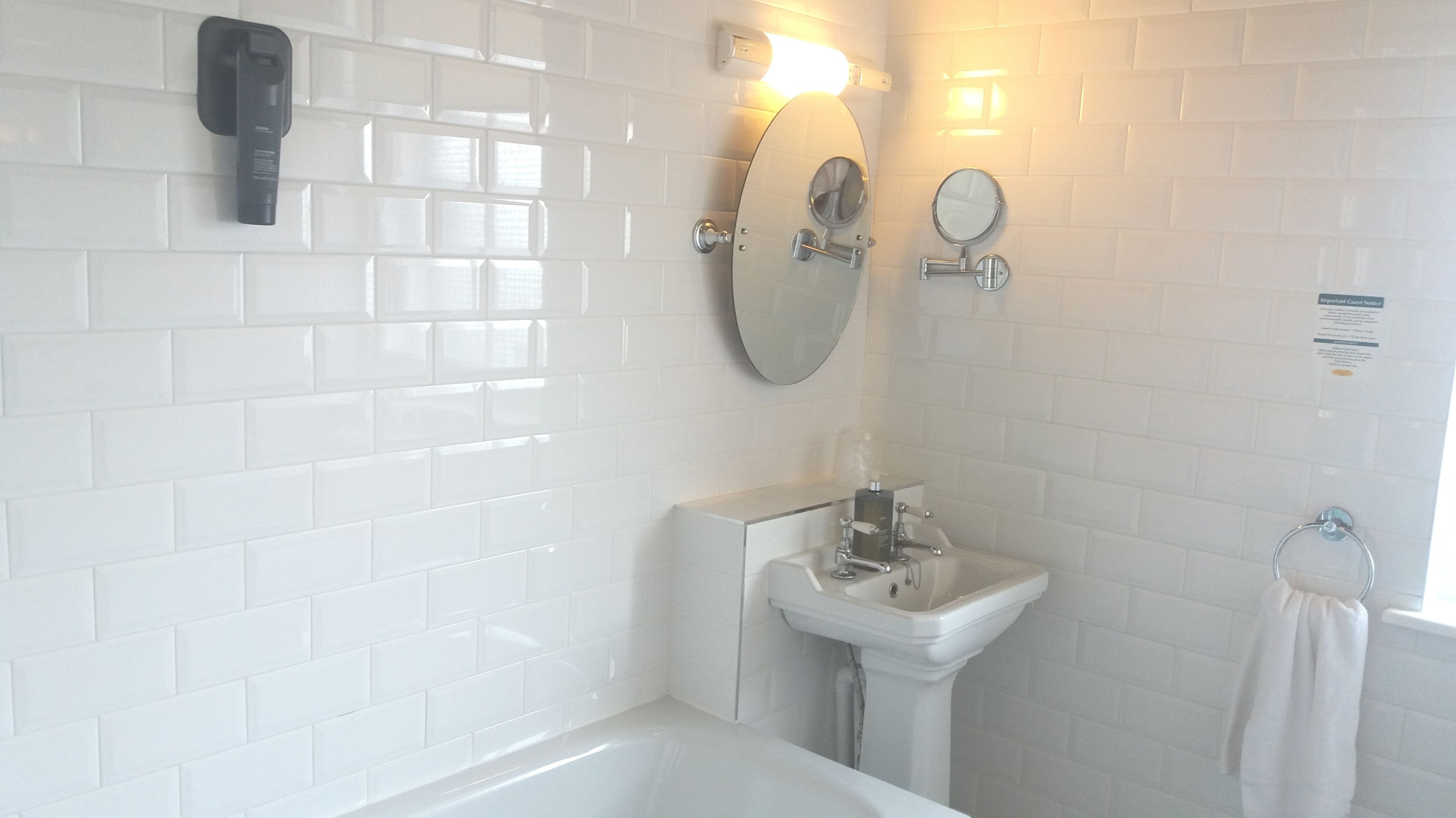 A bathroom with white tiled walls, a sink, a mirror, a handheld showerhead, and a towel rack.