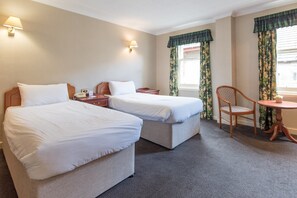 A hotel room with two beds, a small table, a chair, and a window with curtains.