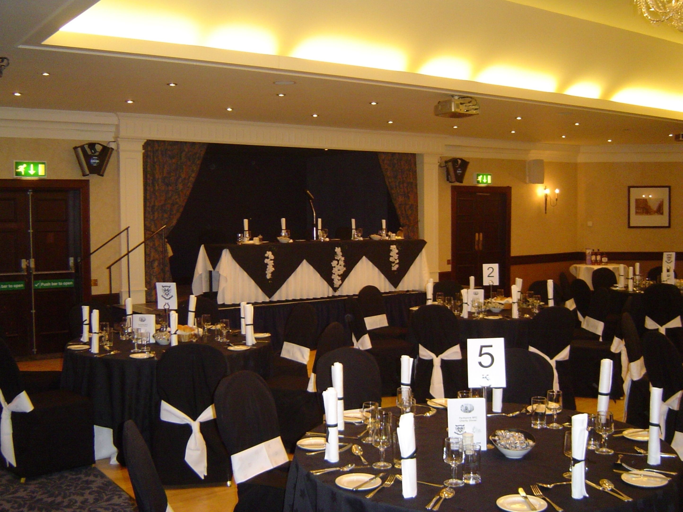 A banquet hall with tables set for a formal event, numbered signs, and a stage with a backdrop.