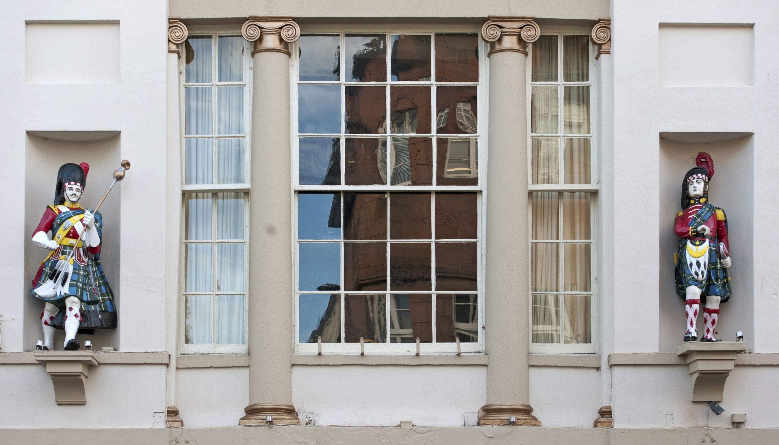 Dos estatuas en la fachada de un edificio, cada una sosteniendo un báculo, situadas a ambos lados de un amplio ventanal que refleja otro edificio.