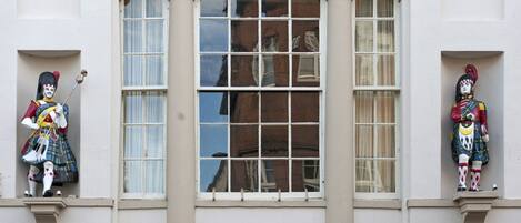 Two statues on a building facade, each holding a staff, positioned on either side of a large window with a reflection of a building behind it.