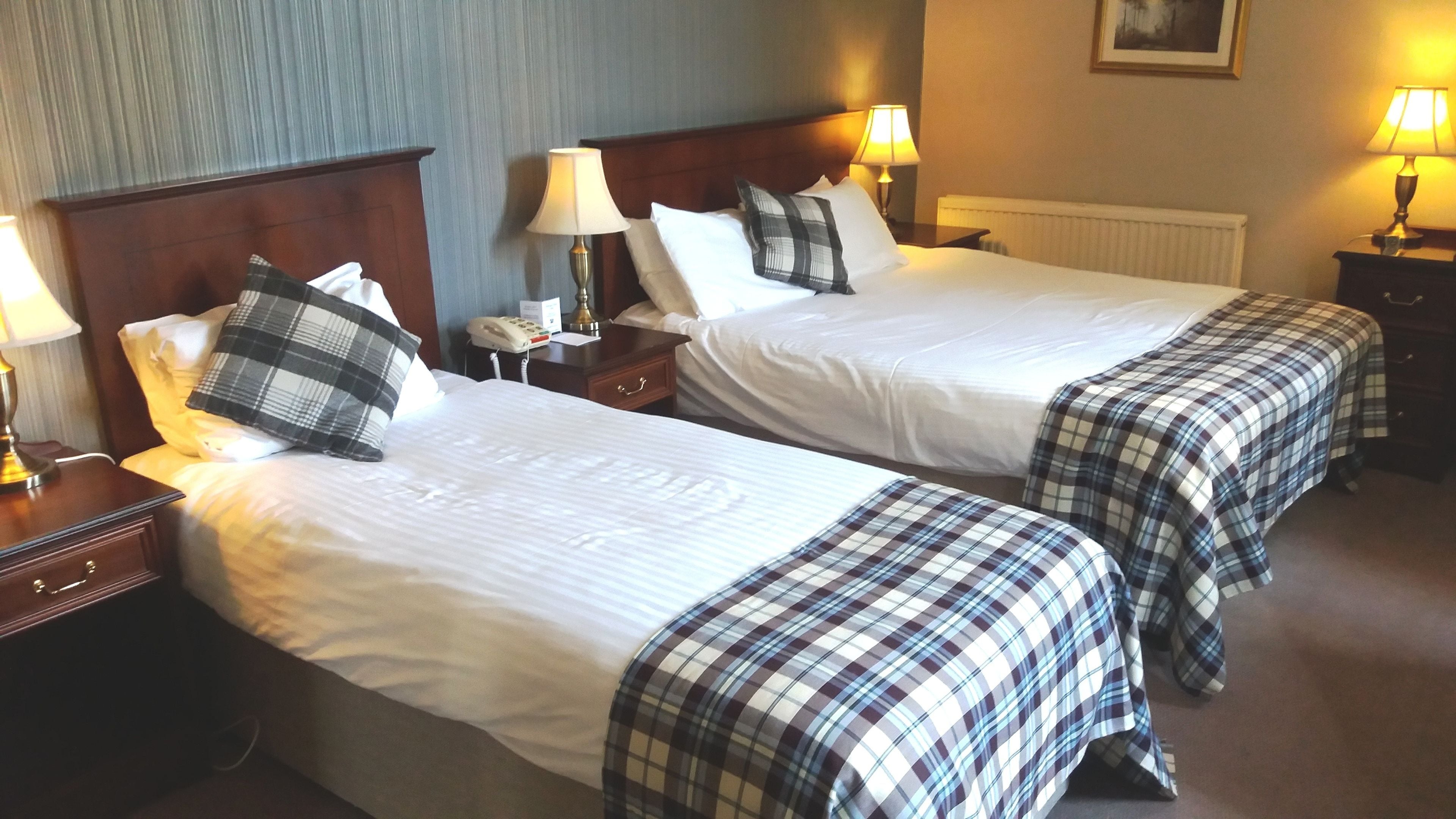 A hotel room with two beds, each with a wooden headboard, white bedding, and plaid bedspreads. There are bedside lamps, a telephone, and a radiator on the wall.
