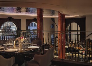 A dining area with a round table set for six, a bar in the background, and a view of the city through large windows.
