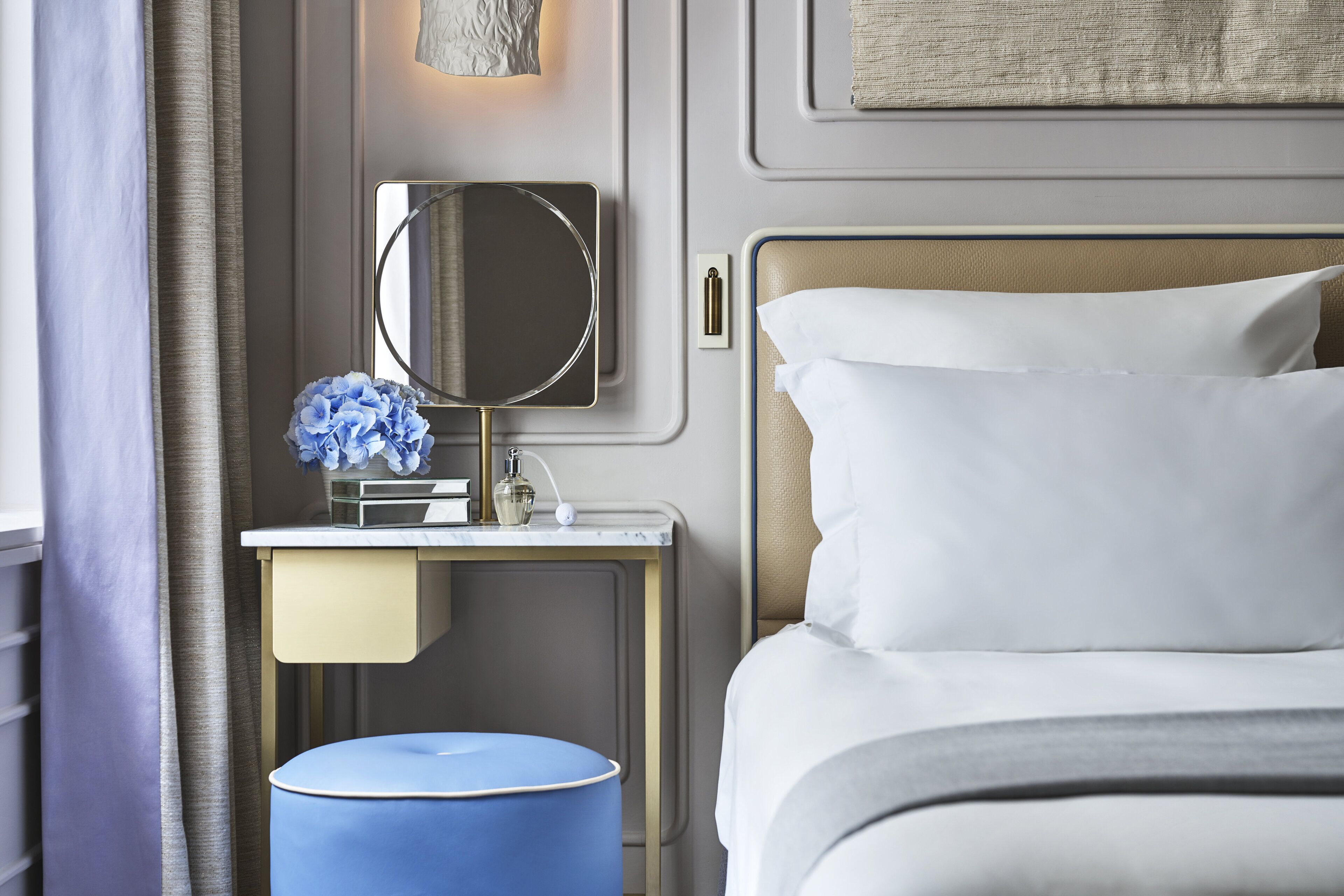 A neatly arranged hotel room with a bed, a bedside table with a mirror, a lamp, and a small blue ottoman.