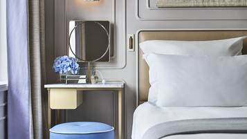 A neatly arranged hotel room with a bed, a bedside table with a mirror, a lamp, and a small blue ottoman.