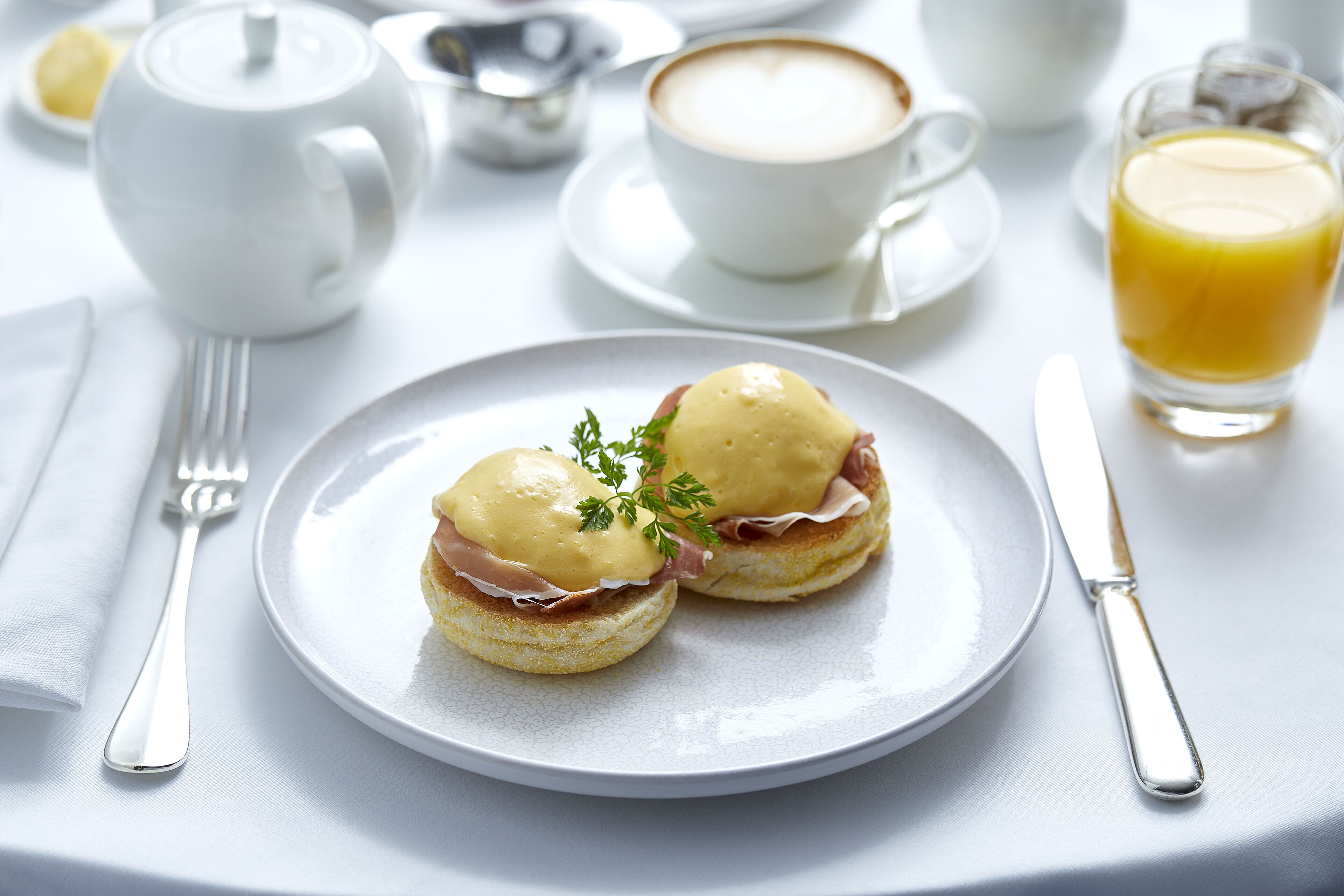 Un desayuno con huevos Benedict en un plato, una taza de café, un calentador de té y un vaso de jugo de naranja.