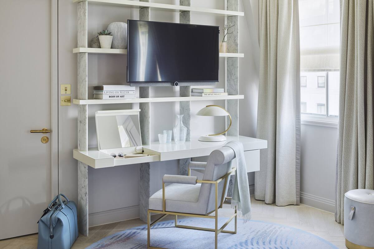 A modern hotel room with a white desk, a flat-screen TV, a comfortable chair, and a small round table.