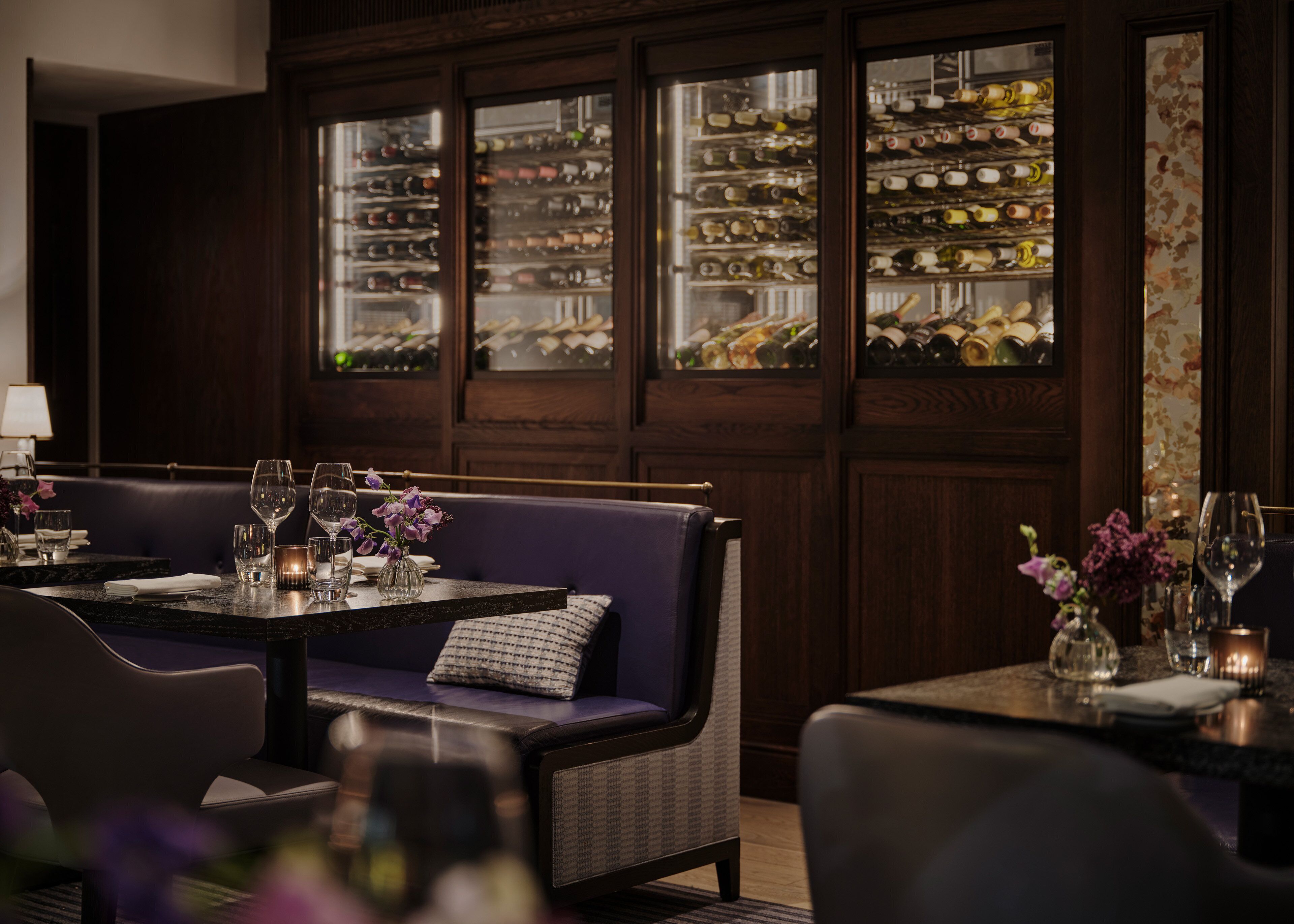 A restaurant with a dark wood-paneled wall, a purple sofa, and a table set with wine glasses and flowers.