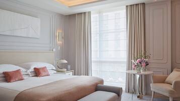 A hotel room with a large bed, a bedside table with a lamp, a chair, a small table with flowers, and a large window with curtains.