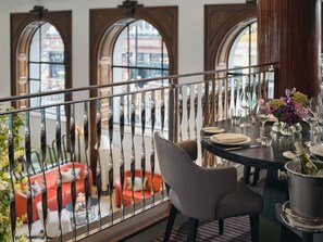 A dining area with a view of the city through arched windows, a balcony railing, and a table set for dining.