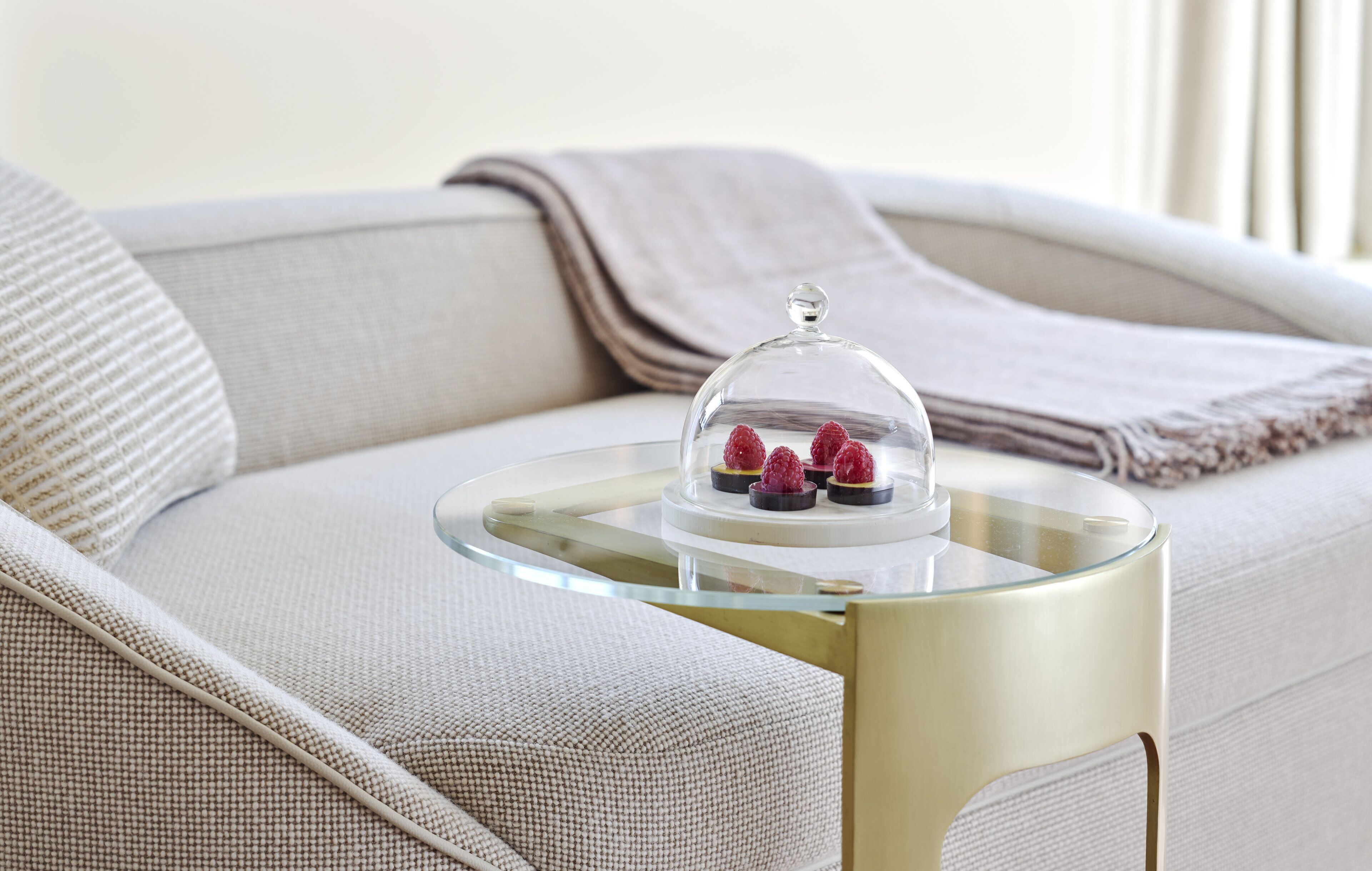 A glass-top table with a clear dome cover holding three raspberries on a beige sofa.