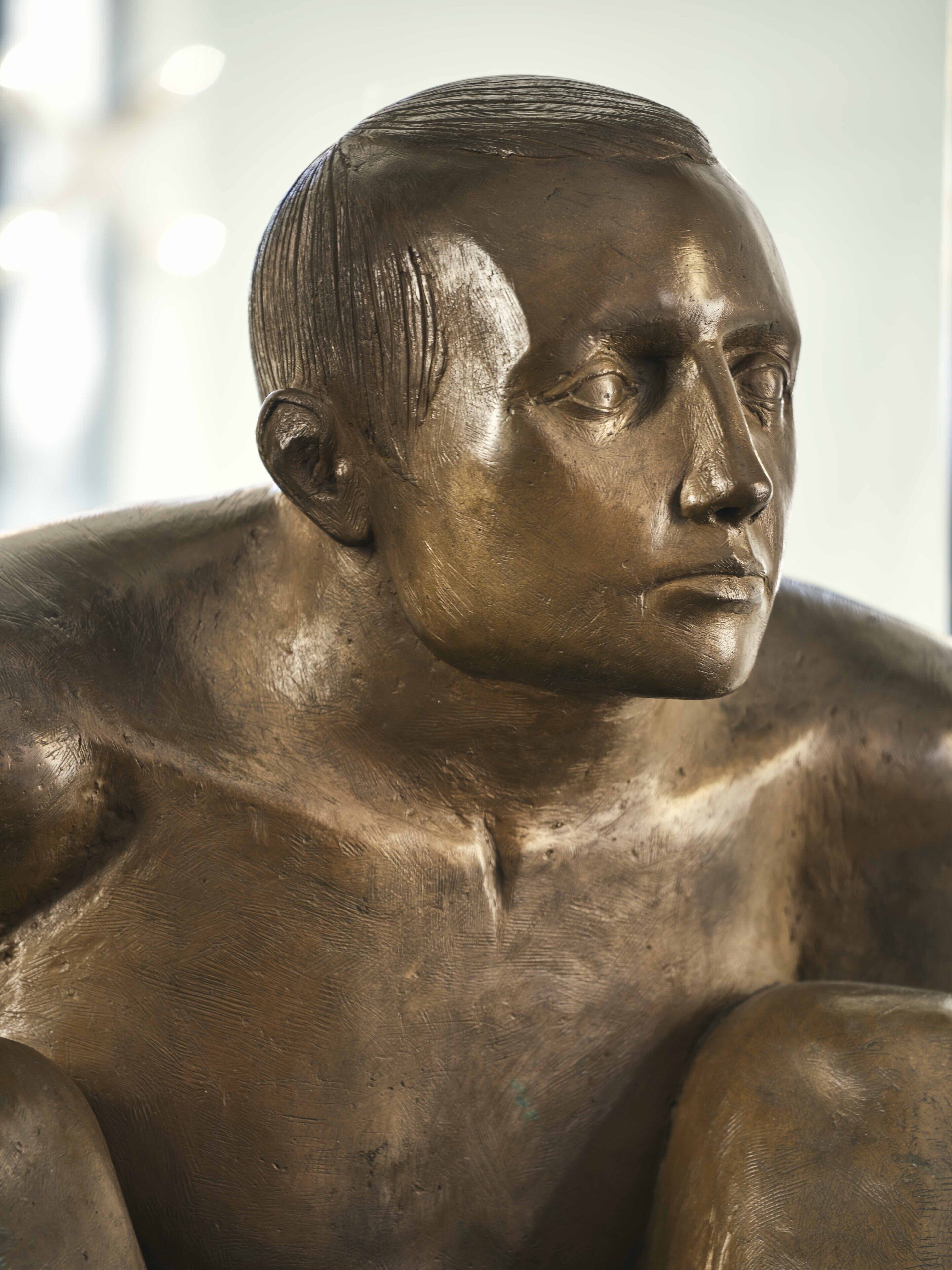 A bronze statue of a seated man with a muscular build, positioned indoors.
