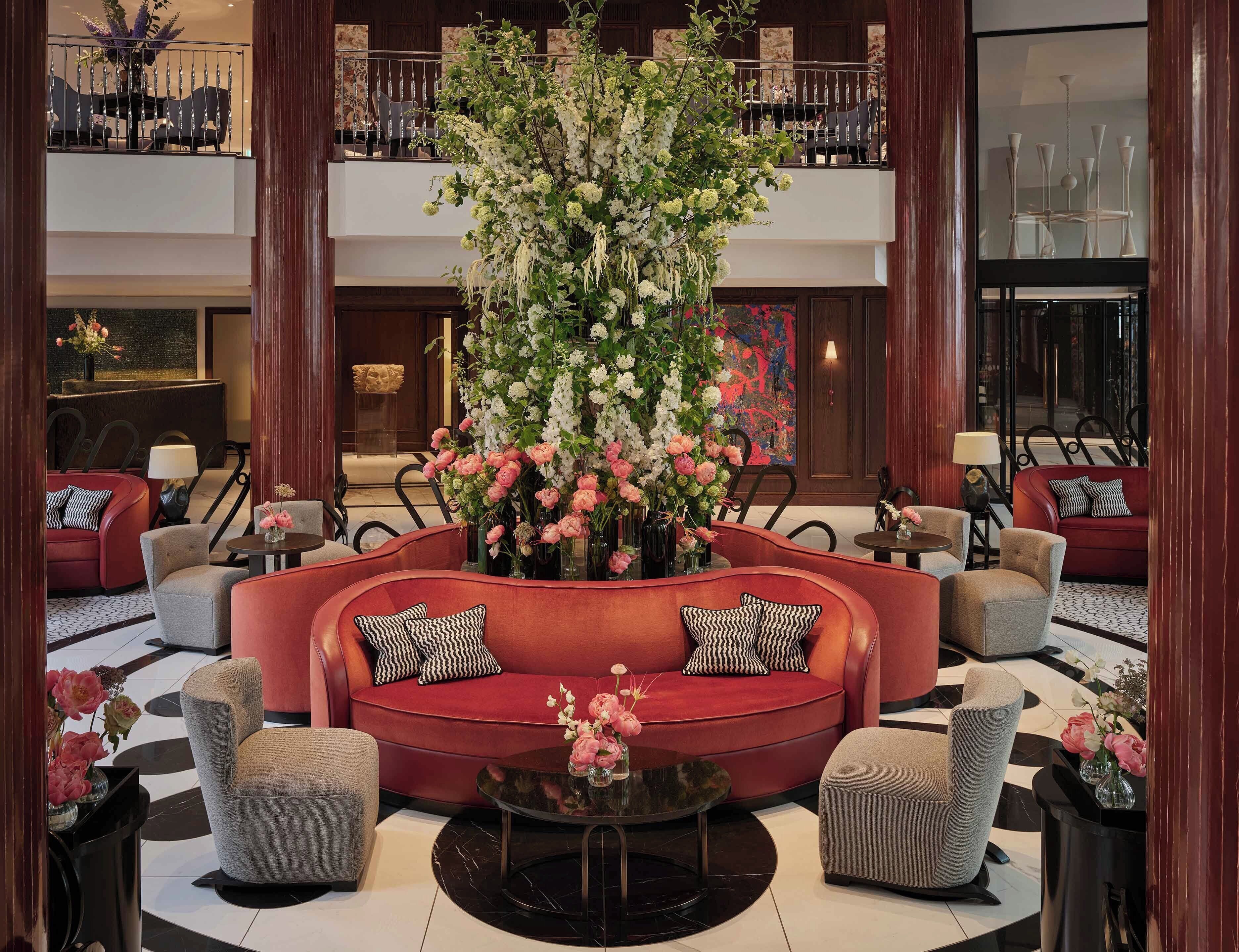 A lobby with a central red sofa, multiple armchairs, a round coffee table, and a large floral arrangement.