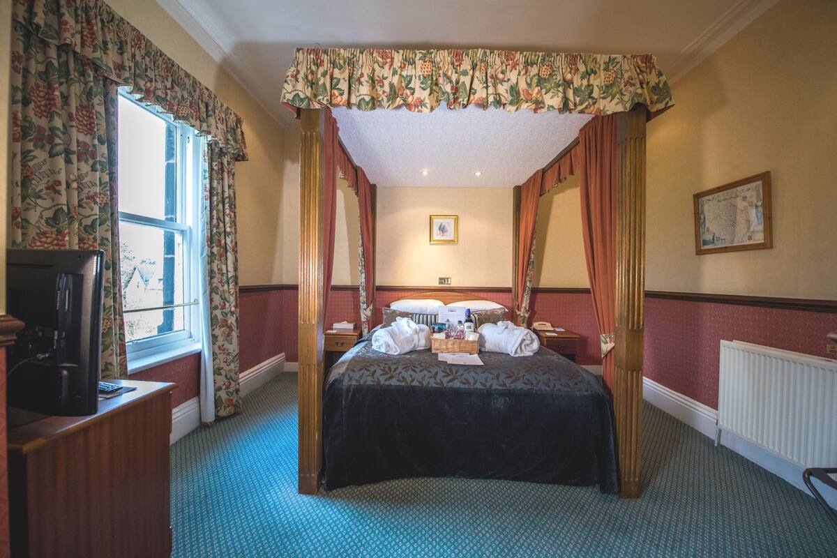 A four-poster canopy bed with a dark bedspread, a bedside table with a lamp, and a window with floral curtains.