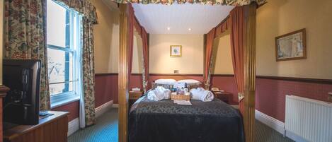 A four-poster canopy bed with a dark bedspread, a bedside table with a lamp, and a window with floral curtains.