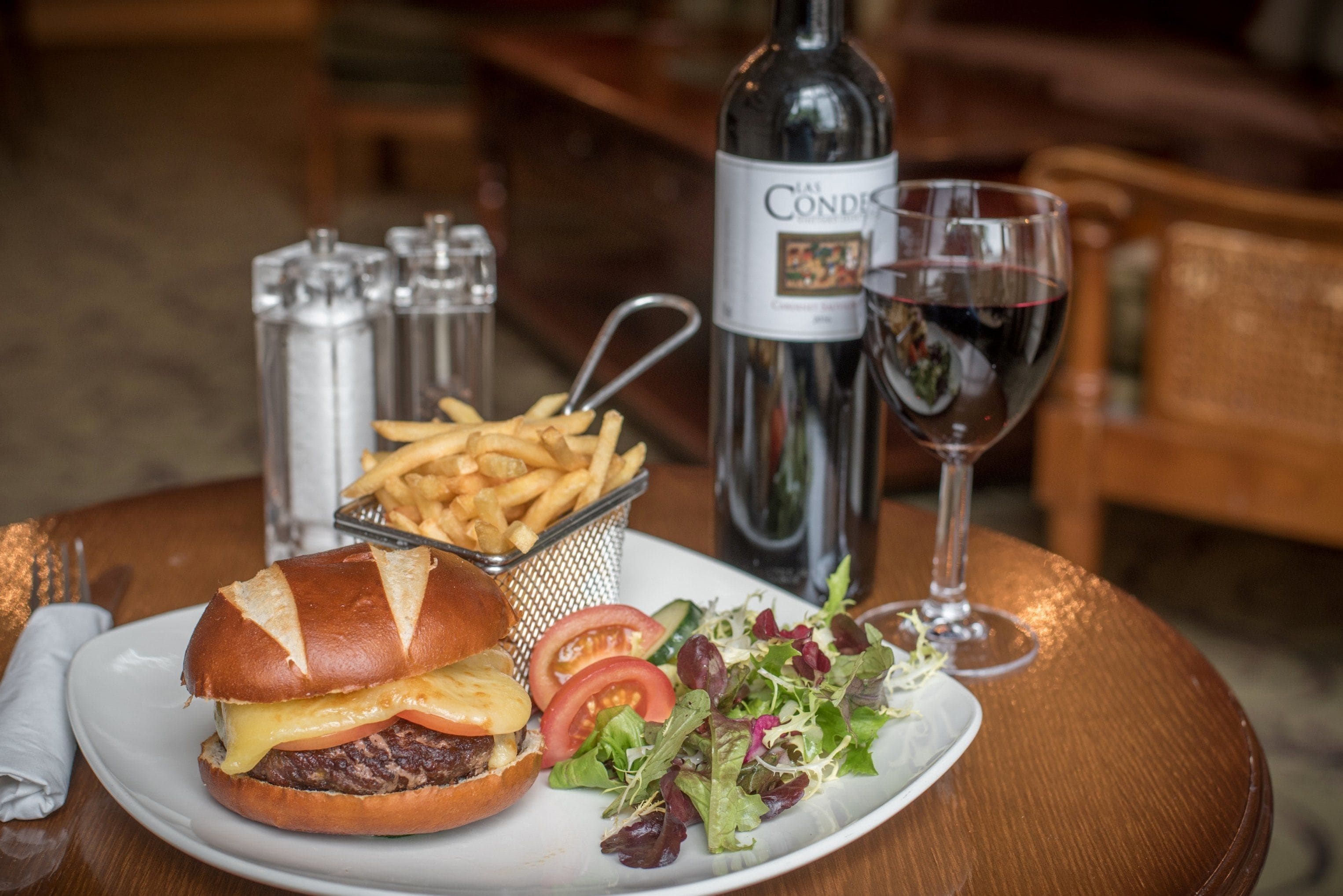 Comida que incluye una hamburguesa, papas fritas, ensalada y una botella de vino sobre una mesa de madera.