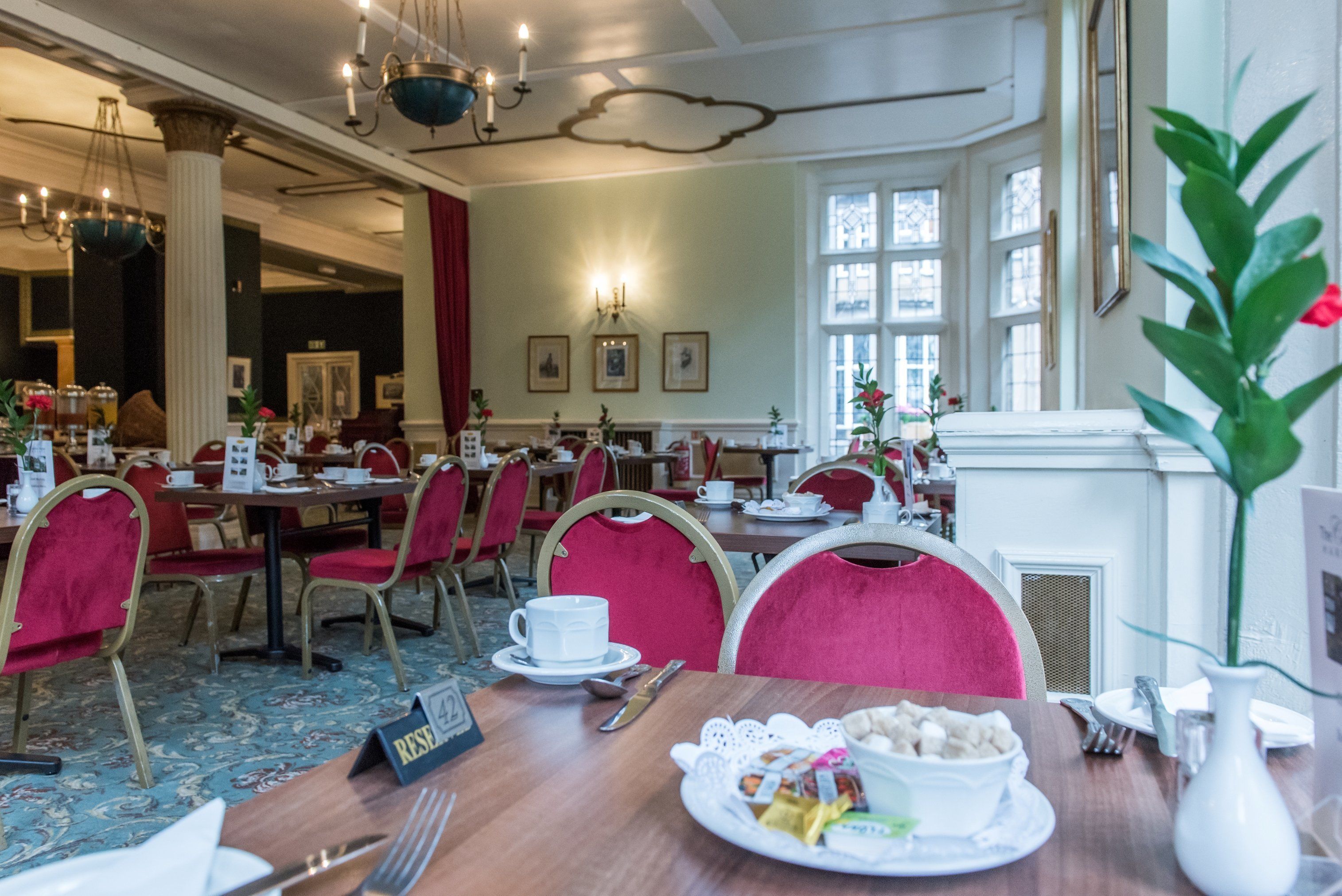 A hotel dining area with tables set for breakfast, red upholstered chairs, and a chandelier.